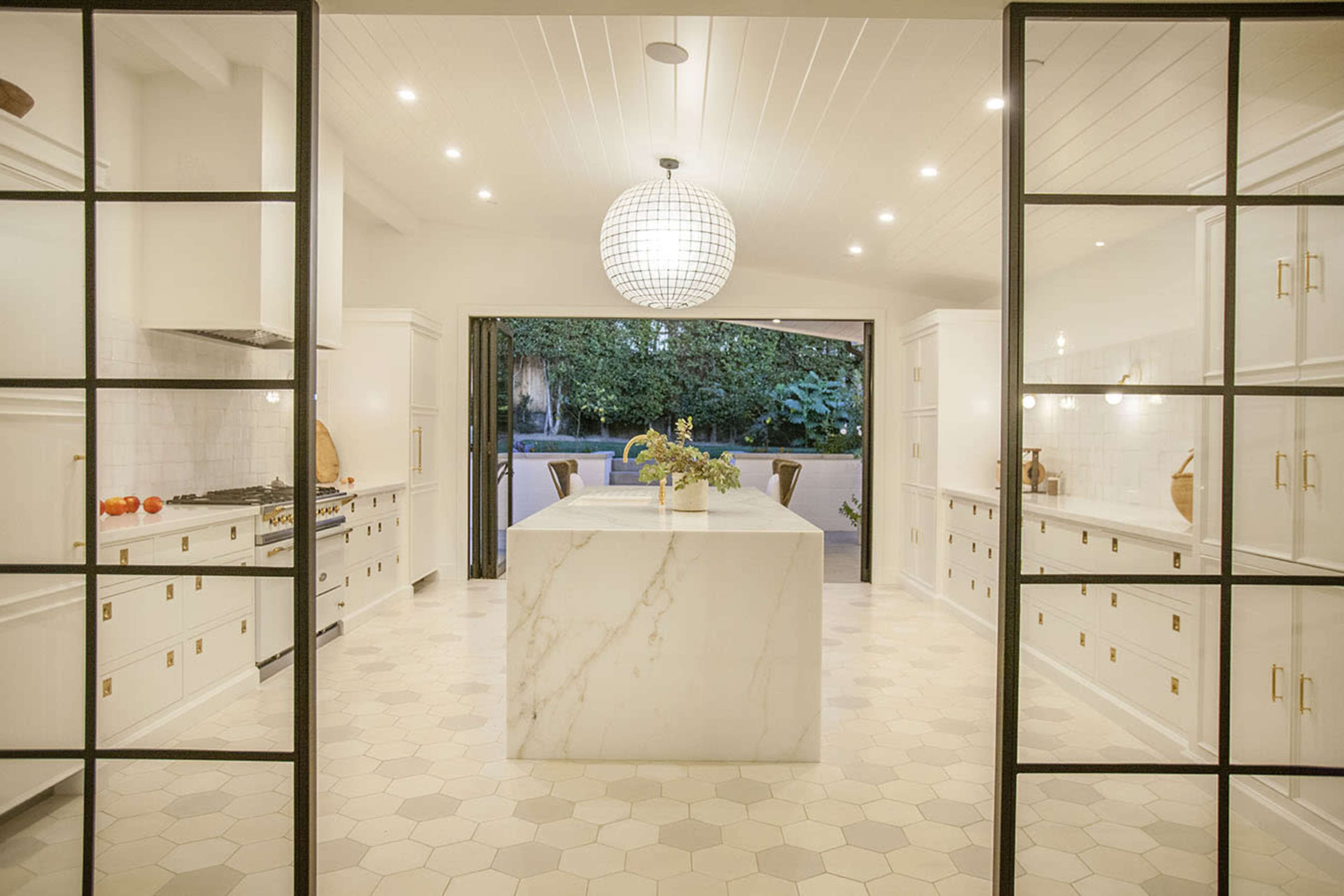 A spacious modern kitchen features a large marble island and white cabinetry with gold handles, illuminated by a round pendant light.