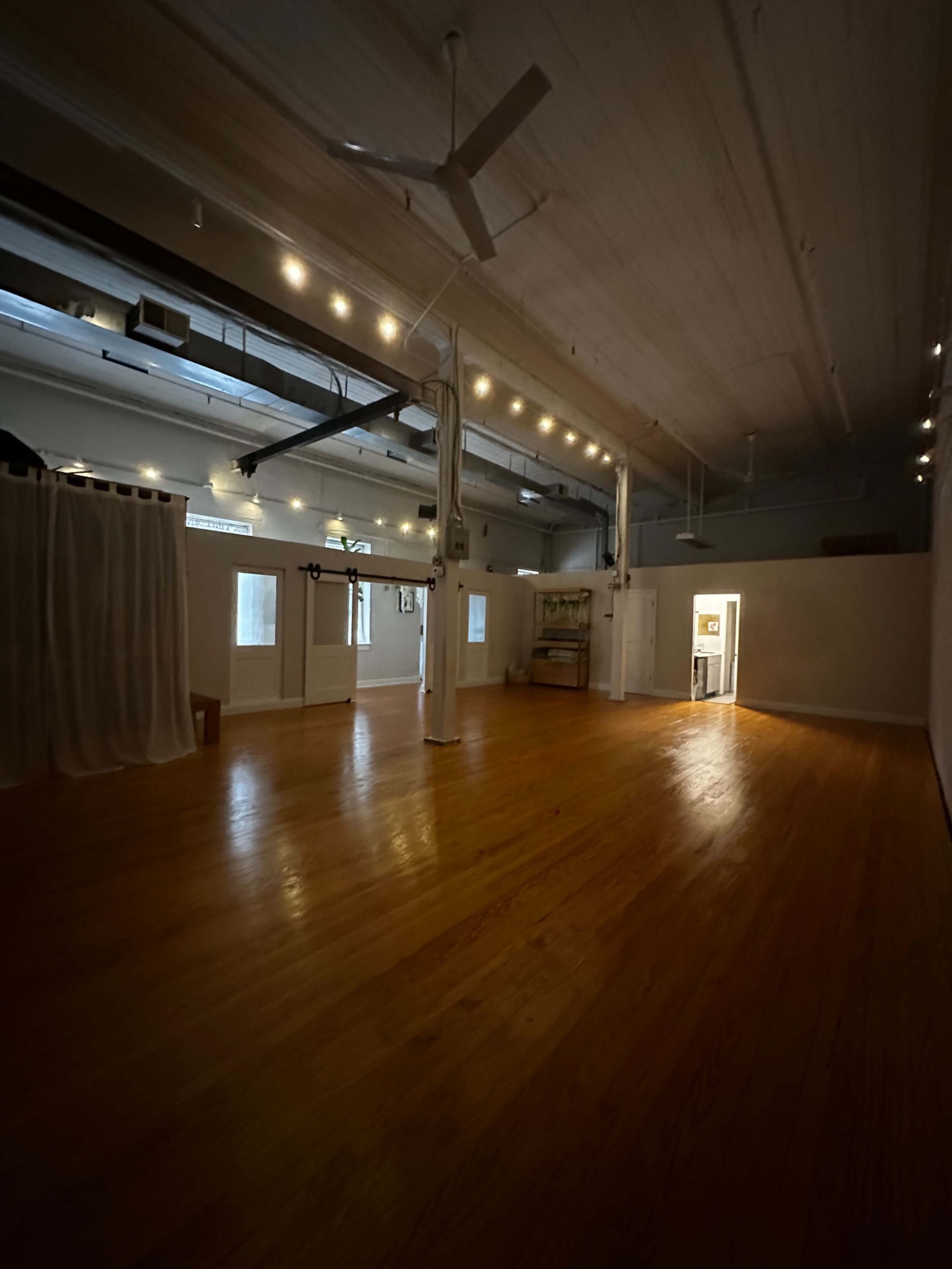 The image shows a spacious, empty indoor room with wooden floors and white walls, illuminated by soft overhead lighting.