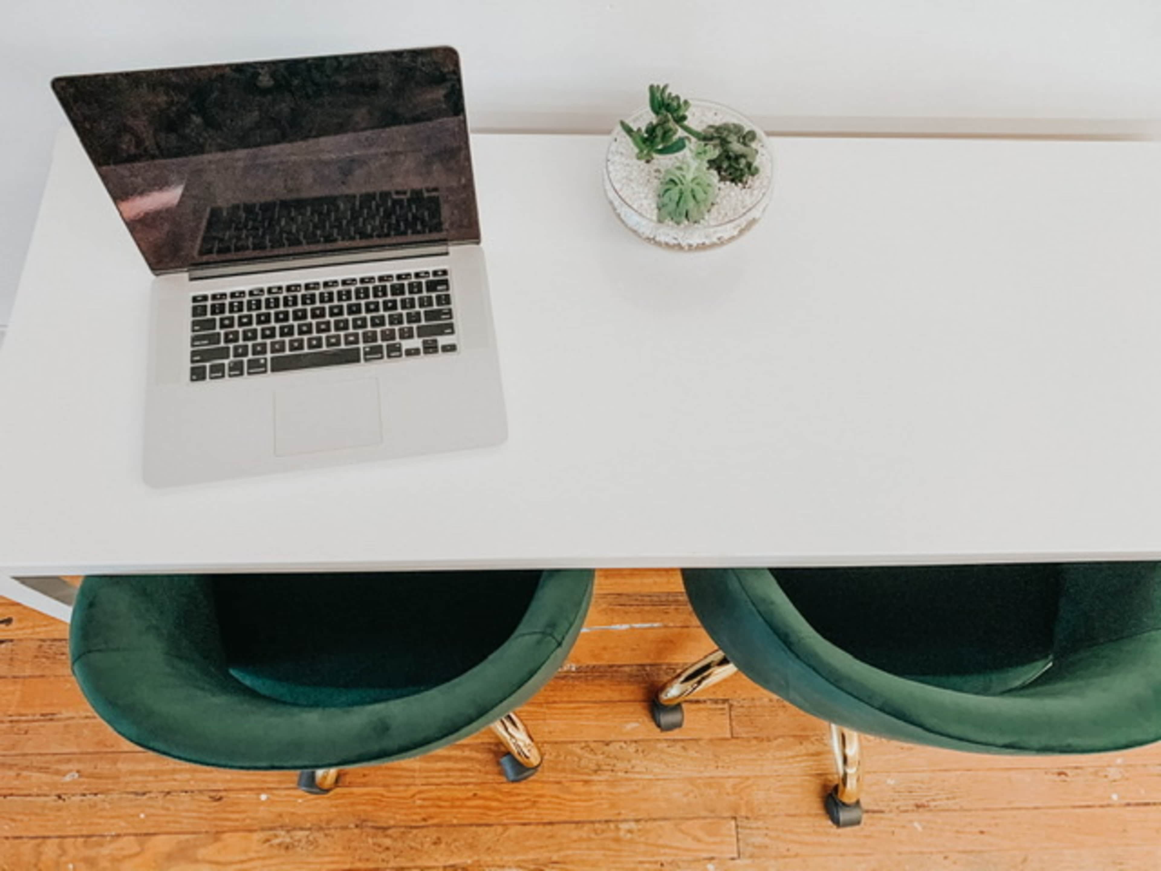A modern workspace features a laptop on a white desk alongside a small planter with succulents and two green chairs.