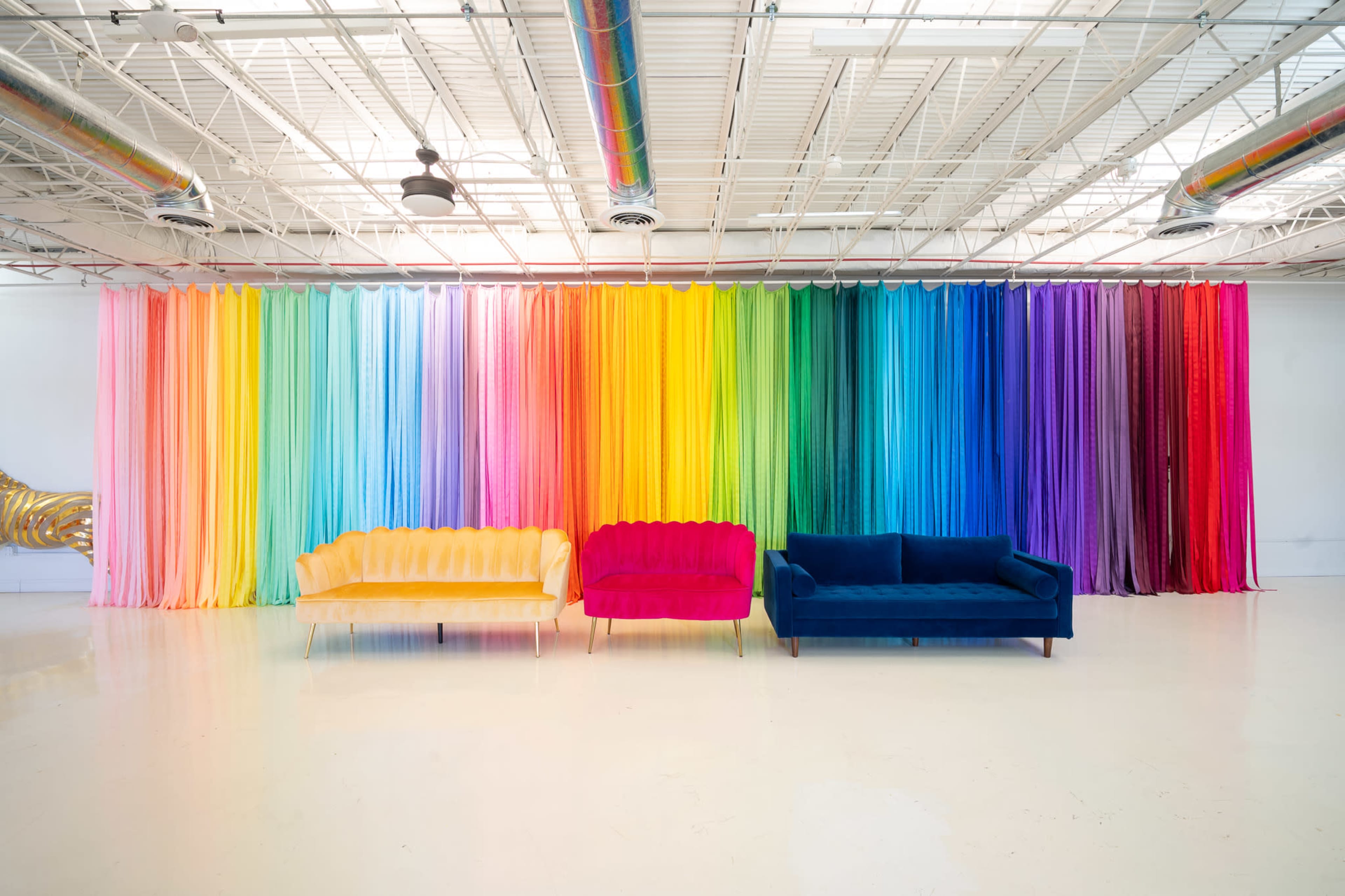 The image features three distinct sofas in front of a gradient rainbow curtain wall, with colors transitioning from yellow to deep purple.