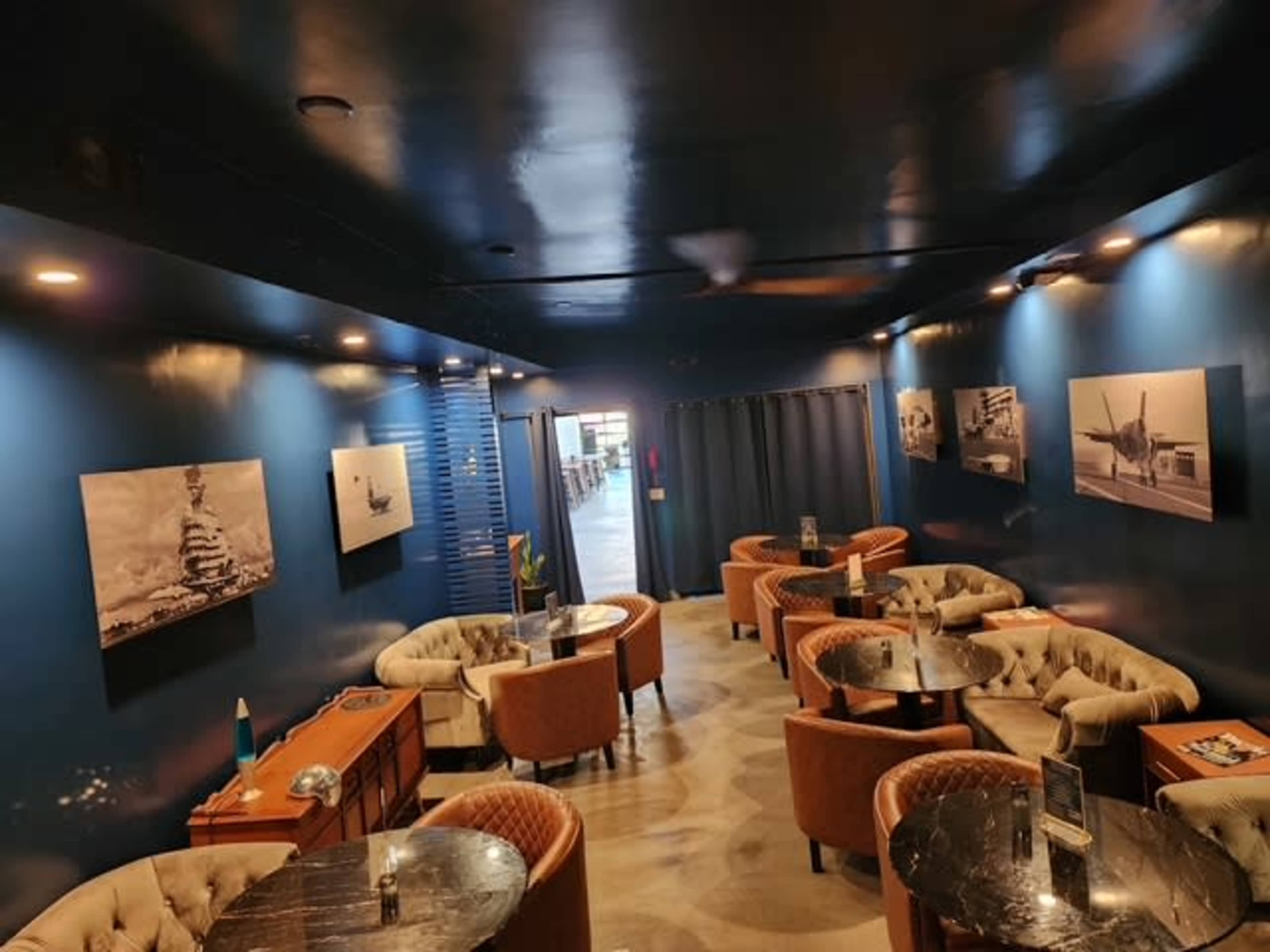 The image depicts a stylish lounge with blue walls, circular tables surrounded by plush armchairs, and black-and-white photographs on the walls.