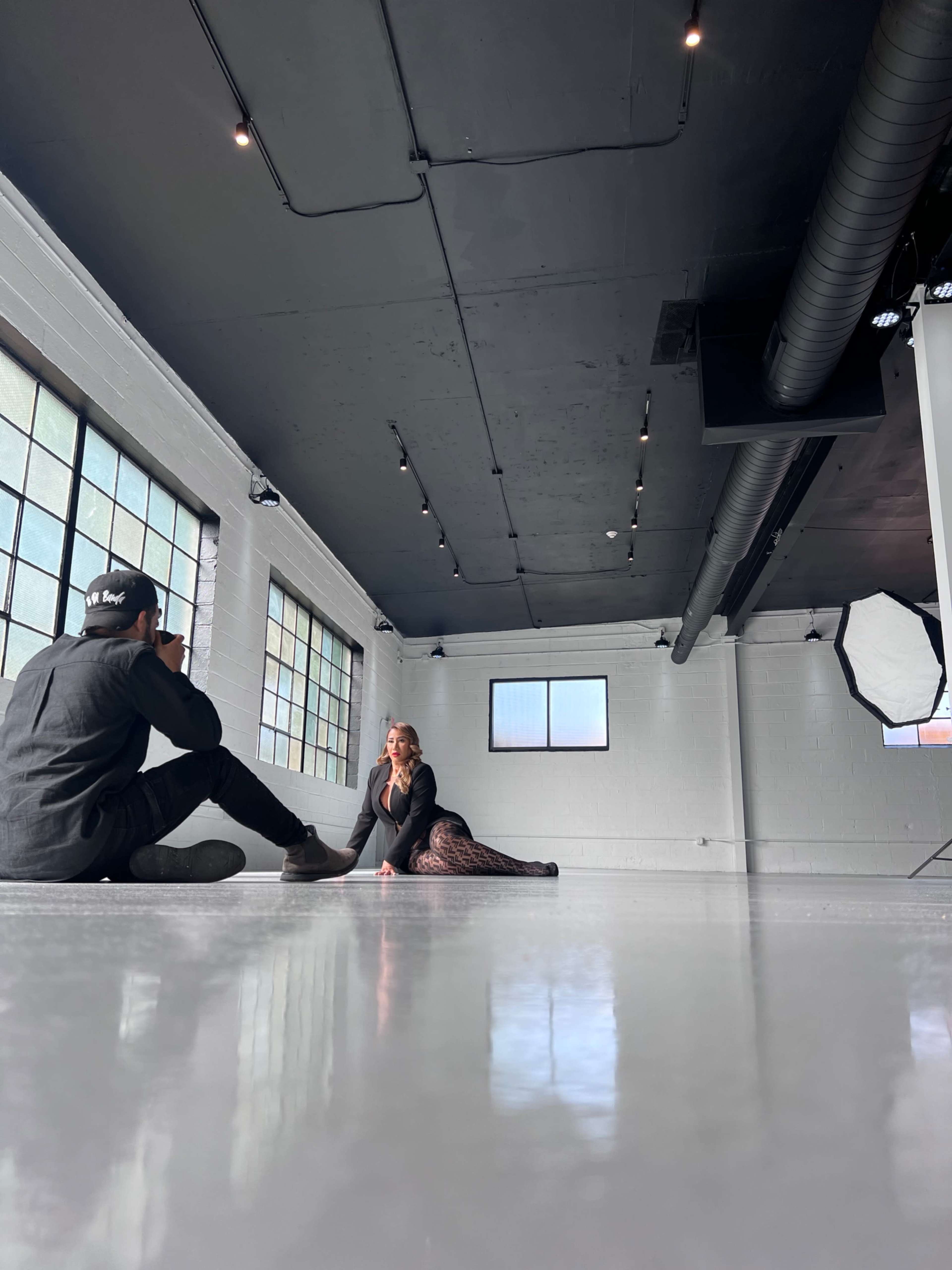 A photographer sits on the floor of a spacious studio while a model poses in front of large windows.