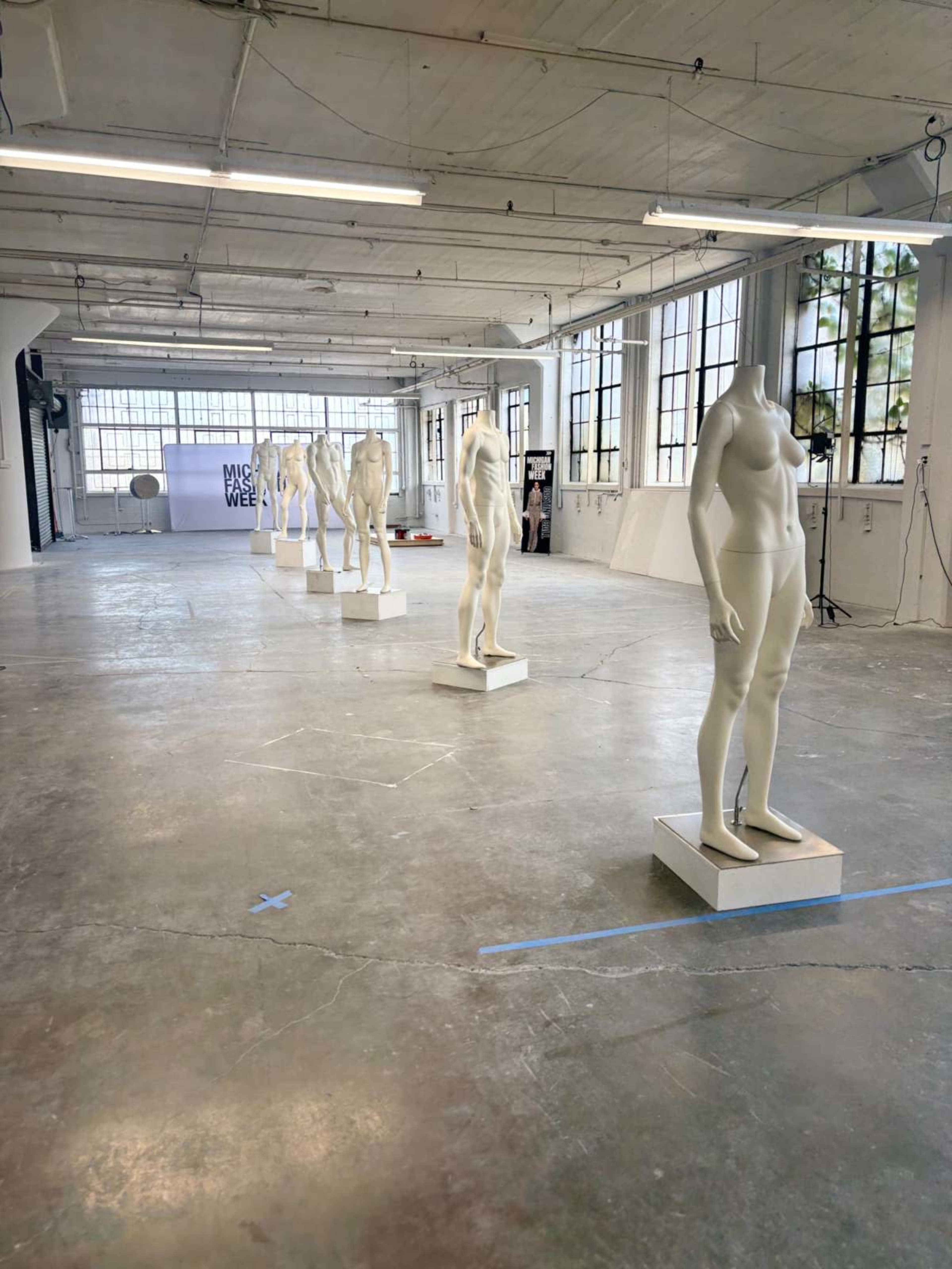 The image shows a spacious industrial room with tall windows and several mannequins displayed on pedestals along the center of the floor.