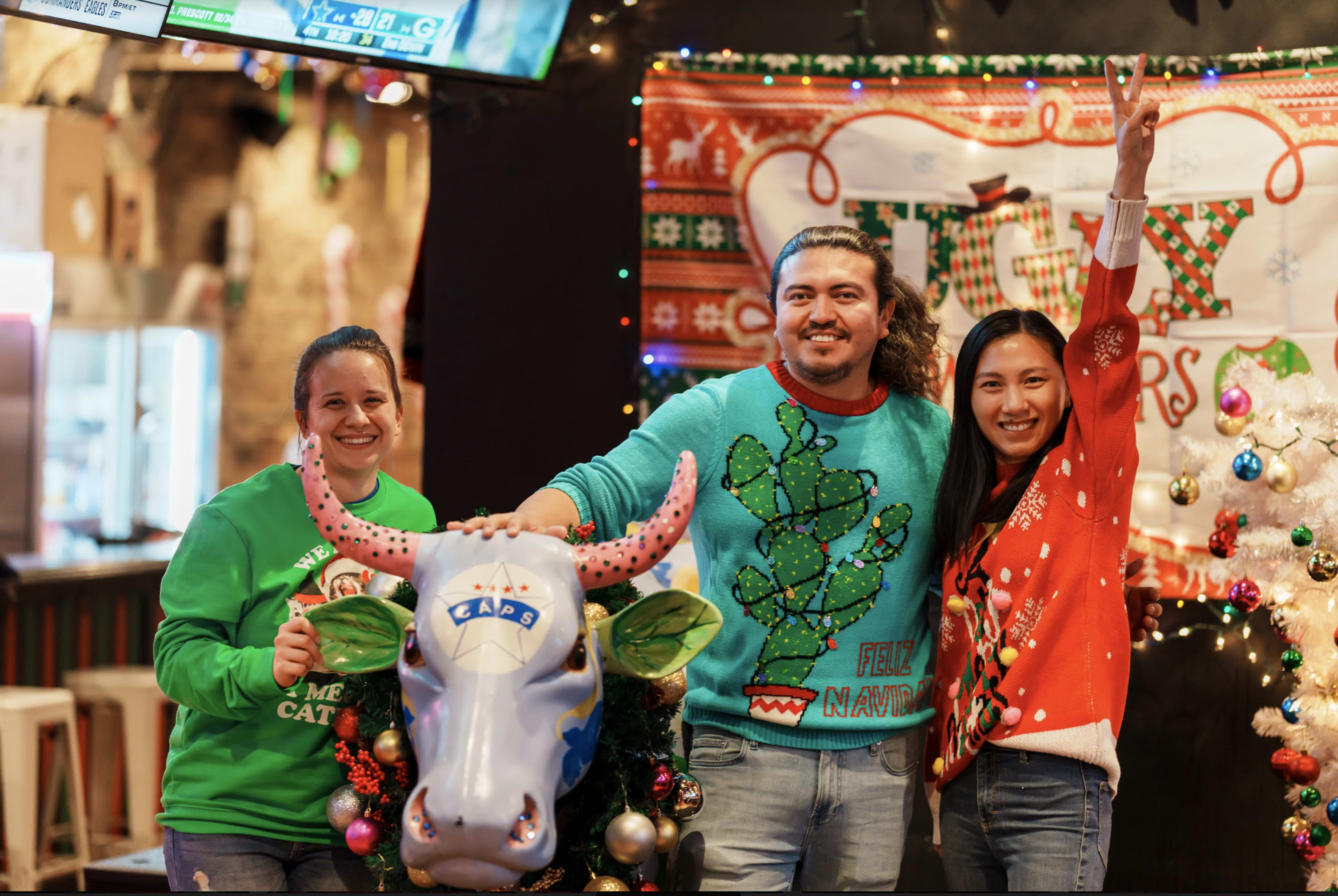Three people pose with a decorated cow statue in a festive setting.