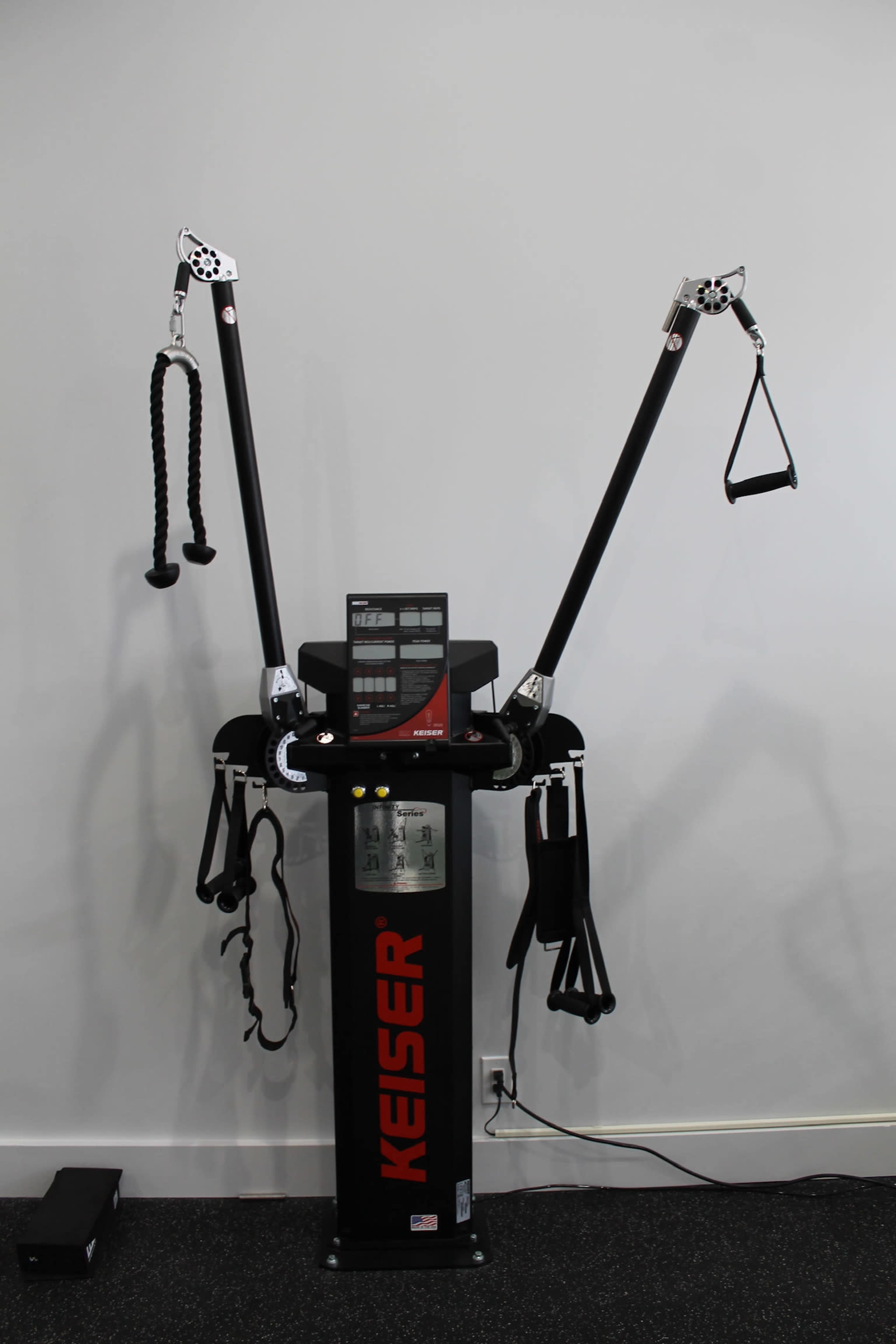 A KEISER strength training machine with dual arms and various attachments set against a plain wall.