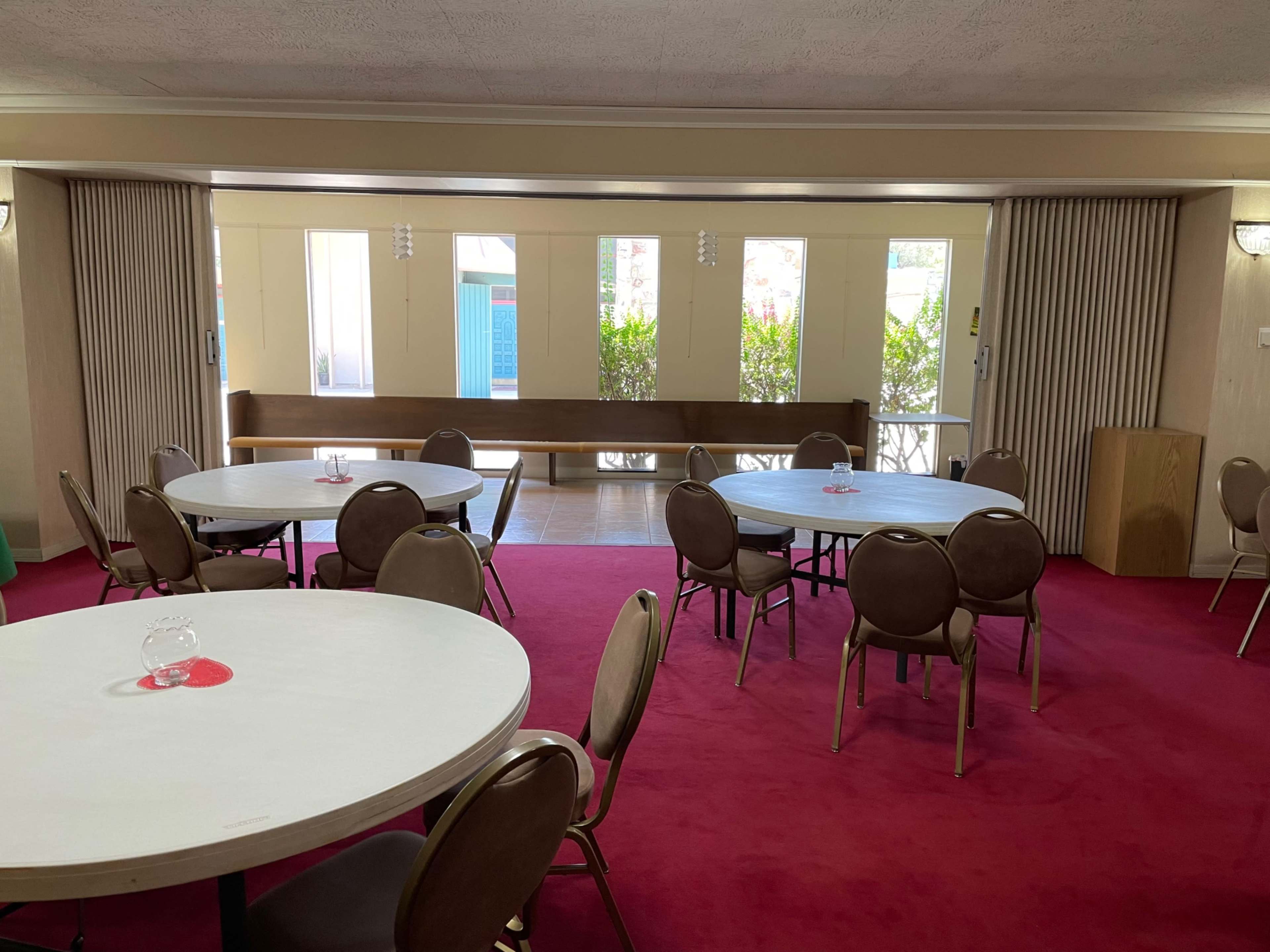 A spacious room with round tables arranged on a red carpet and large windows letting in natural light.