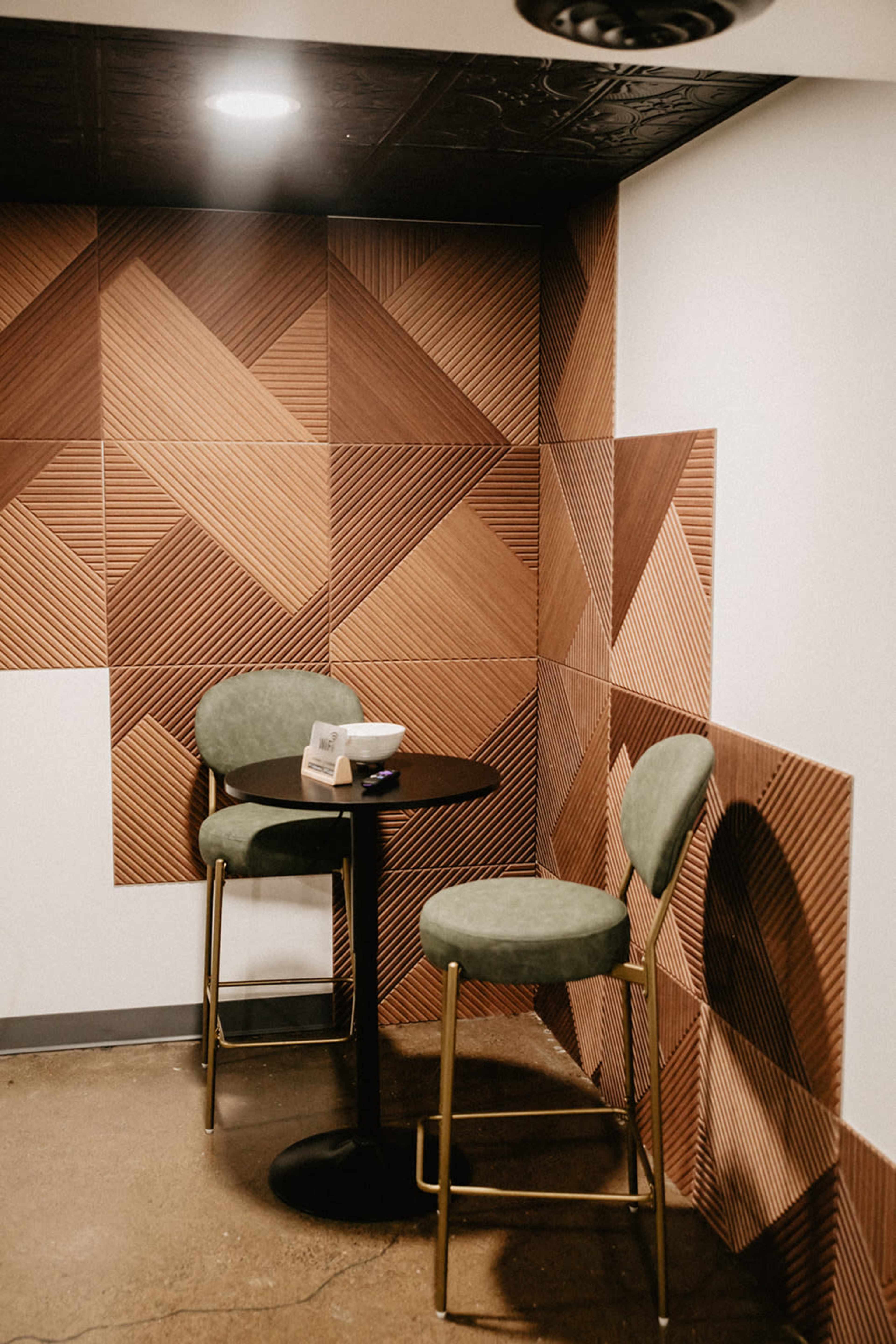 The image shows a small corner of a room featuring a round black table and two green bar stools against a textured wall of geometric patterns in shades of brown.