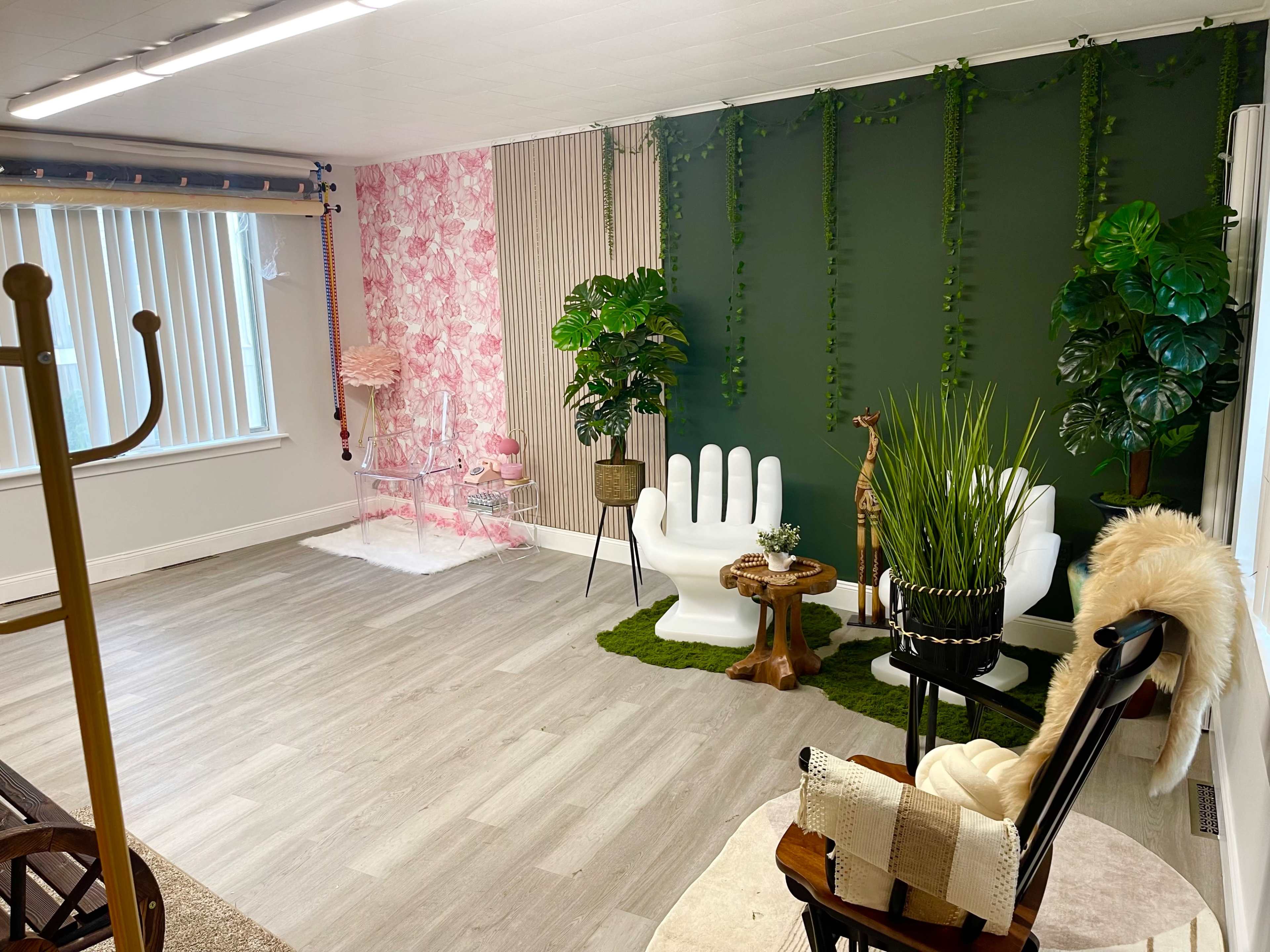 A brightly lit room with a green accent wall, decorative plants, a white hand-shaped chair, and a small wooden table on a light gray floor.