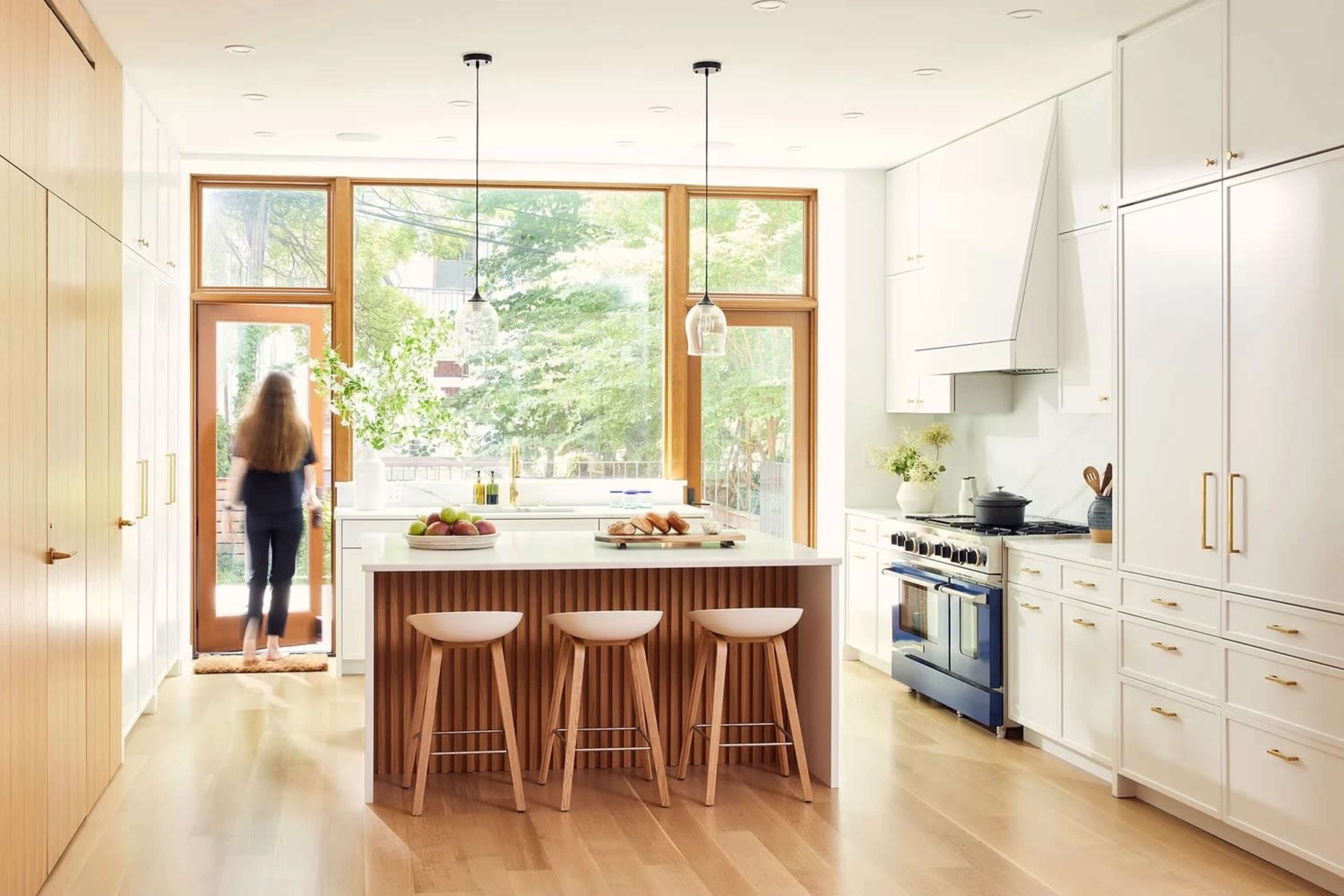A modern kitchen features a large island with three stools, white cabinetry, and large windows allowing natural light to illuminate the space as a person walks towards the entrance.