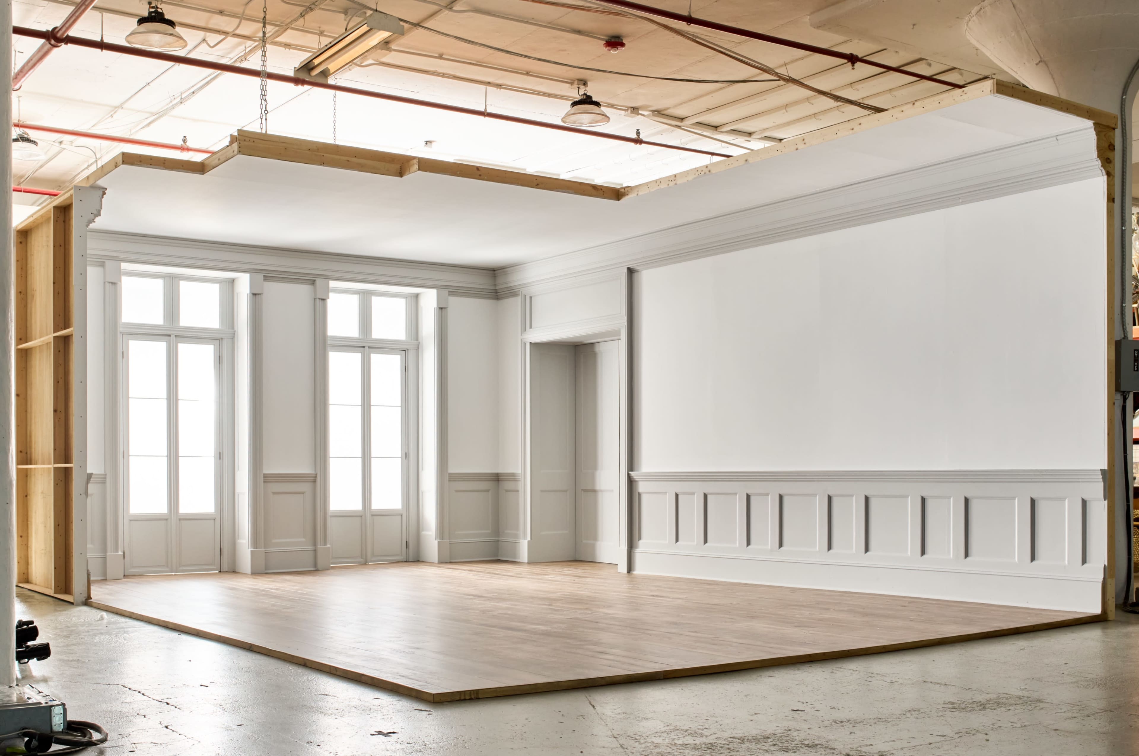 The image shows an empty, well-lit interior space with plain white walls, large windows, and a wooden floor.