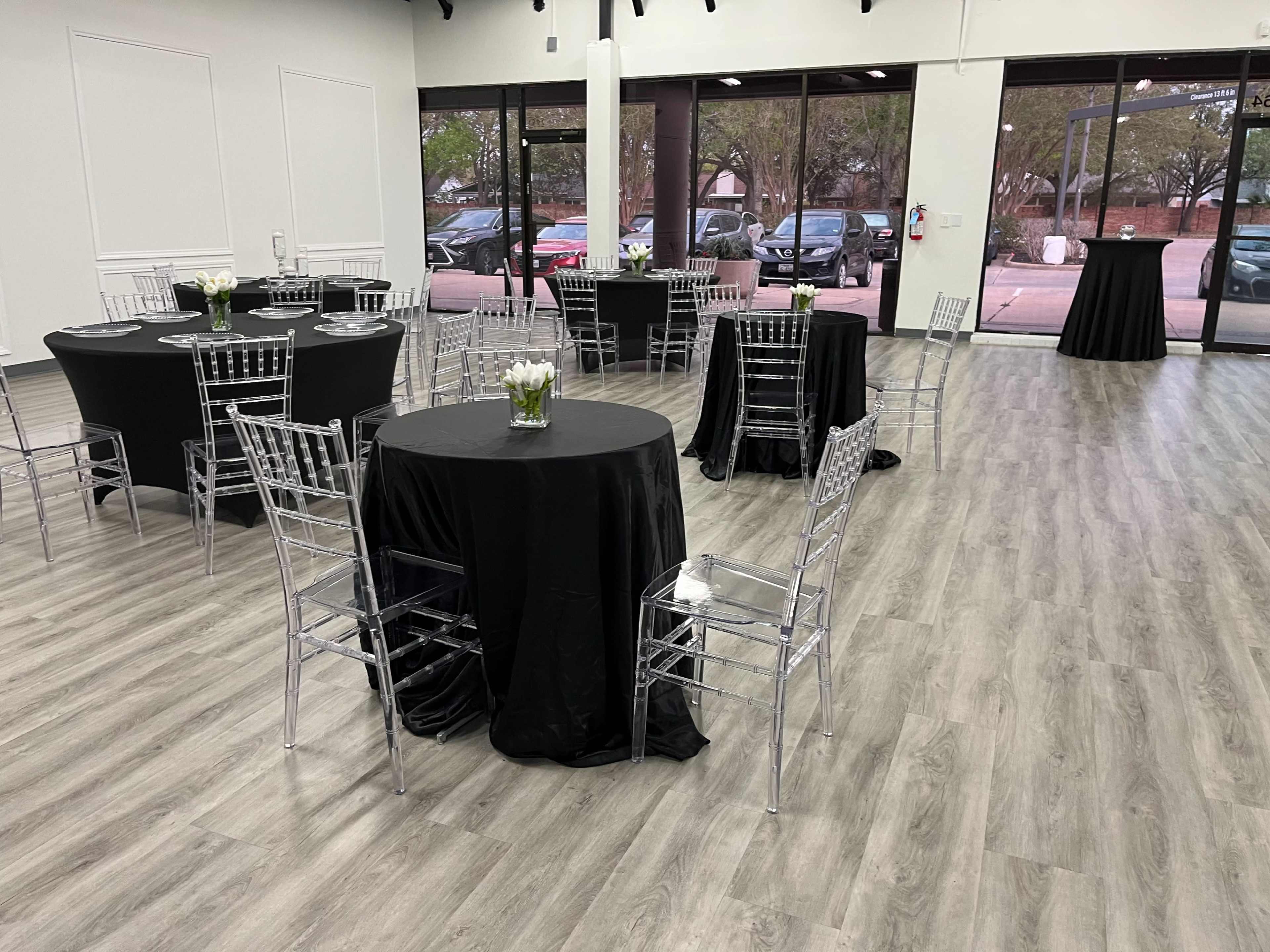 The image shows an event space with several black-covered round tables, each surrounded by clear chiavari chairs, arranged in a well-lit area with large windows.