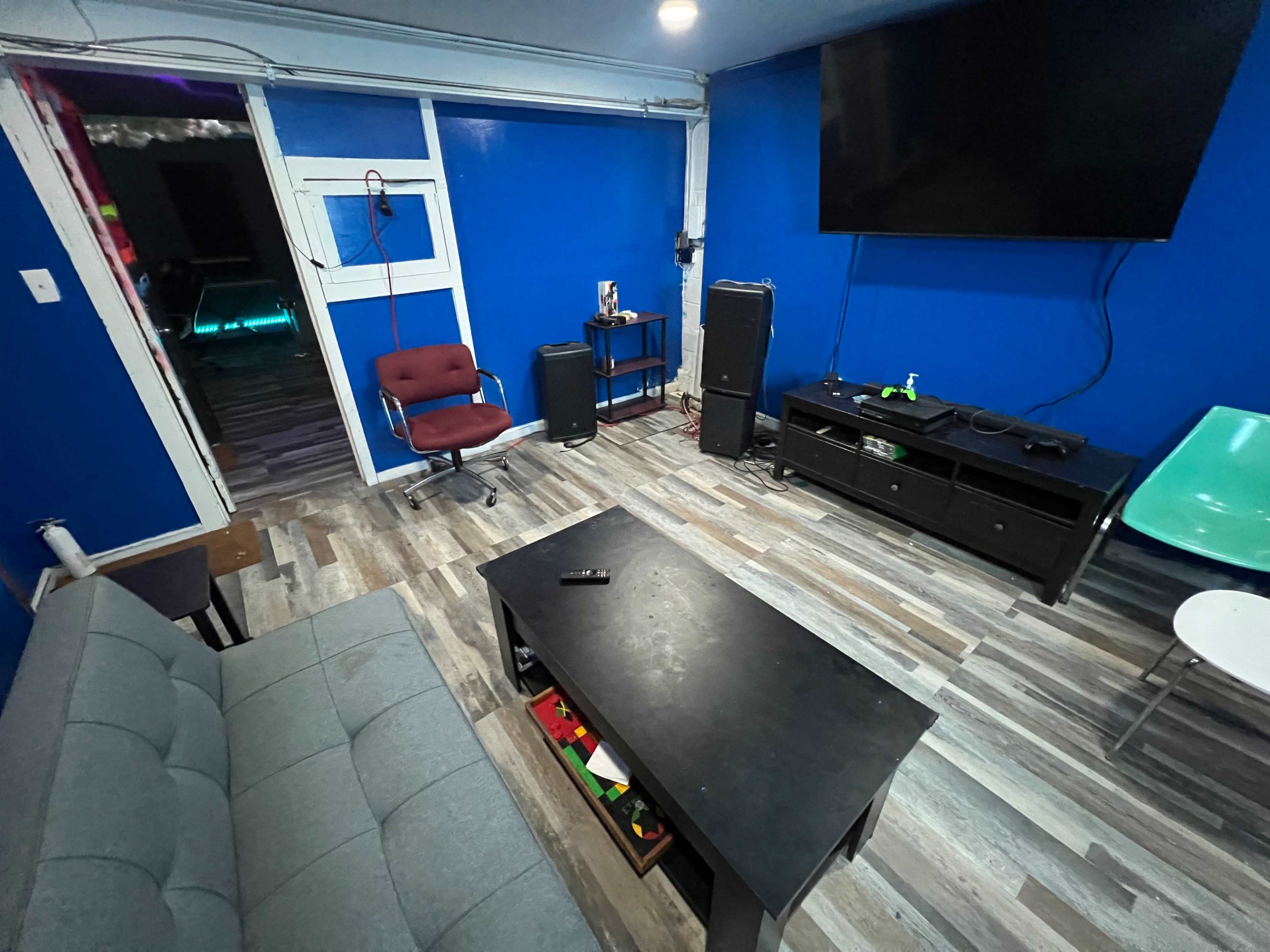 A room with a blue wall, a gray couch, a black coffee table, a television mounted on the wall, and various pieces of furniture, including a red chair and a white chair.