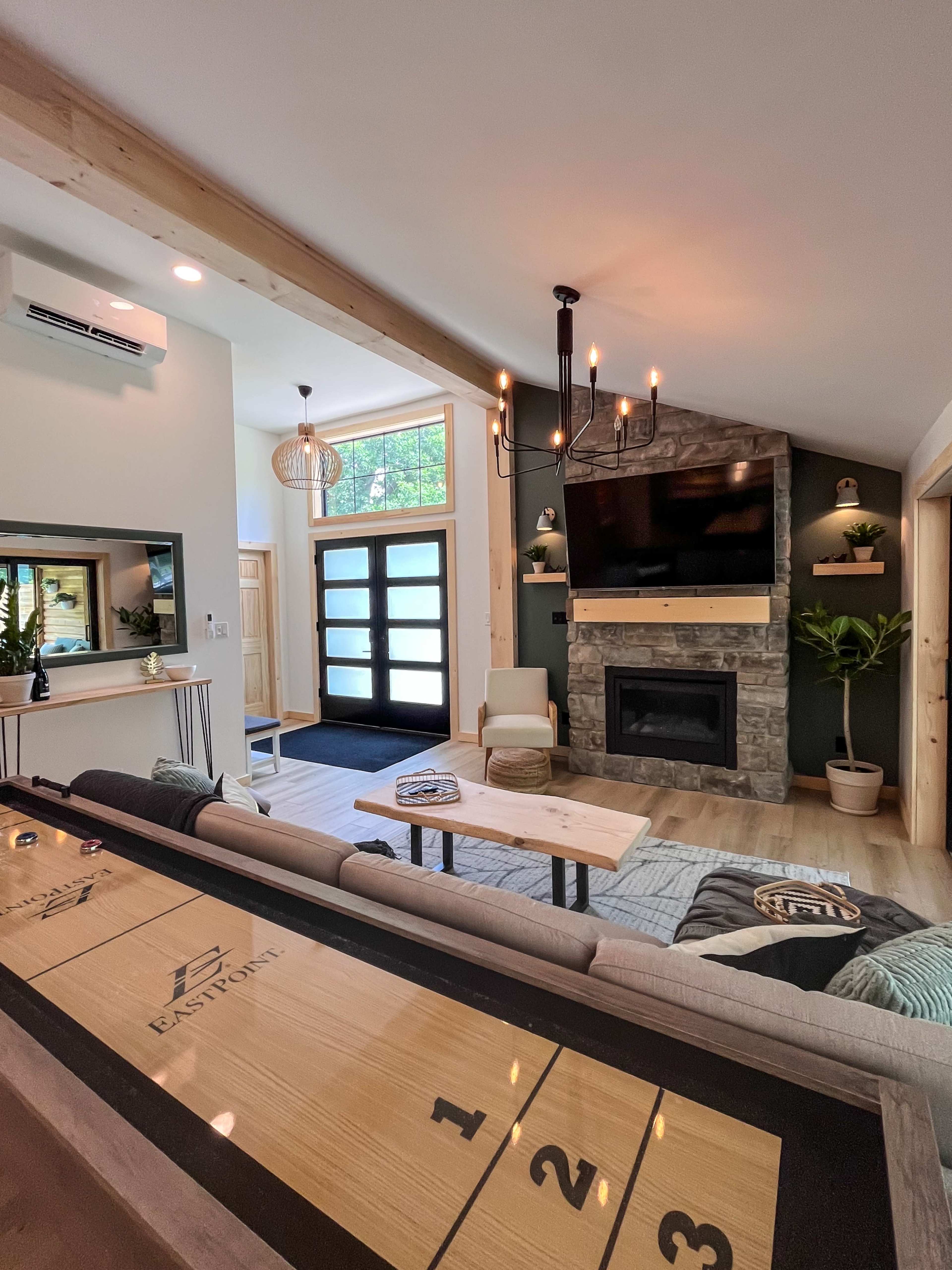 A living room featuring a stone fireplace, a mounted television, a shuffleboard table, and large windows with natural light.