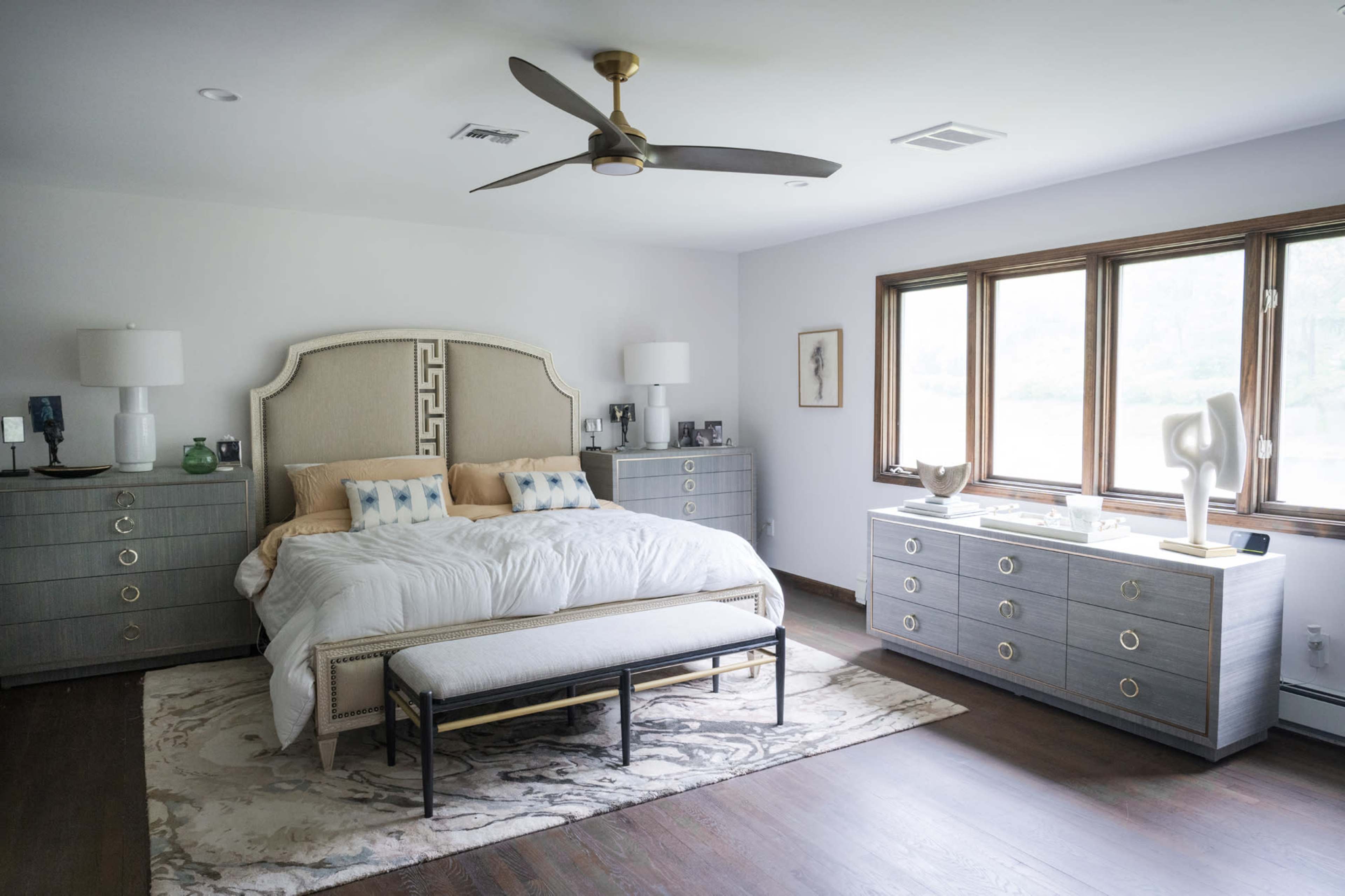 The image shows a modern bedroom featuring a large bed with decorative pillows, dual bedside tables, a central bench, and large windows allowing natural light to fill the space.