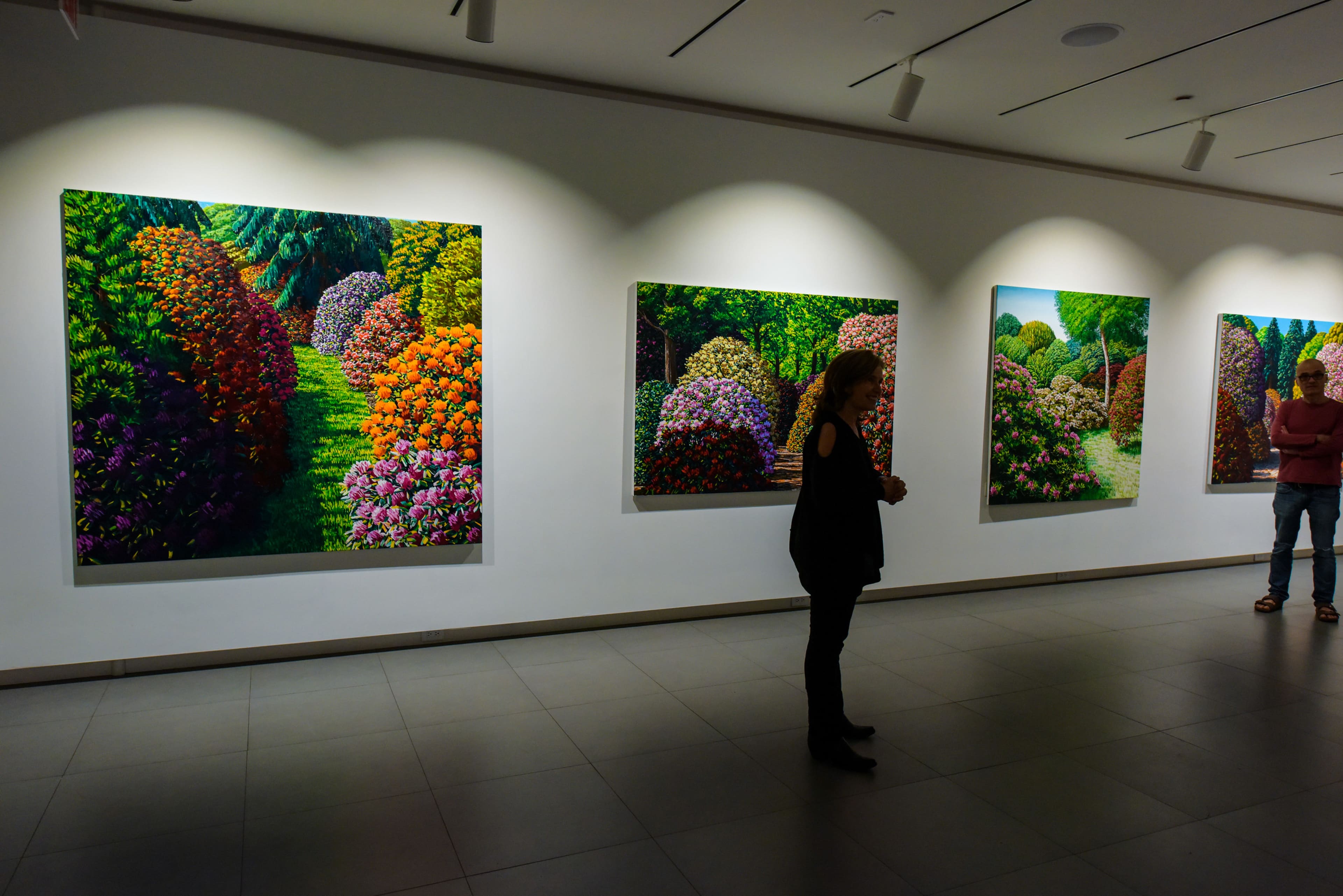A woman stands in an art gallery in front of large, colorful landscape paintings depicting lush gardens.