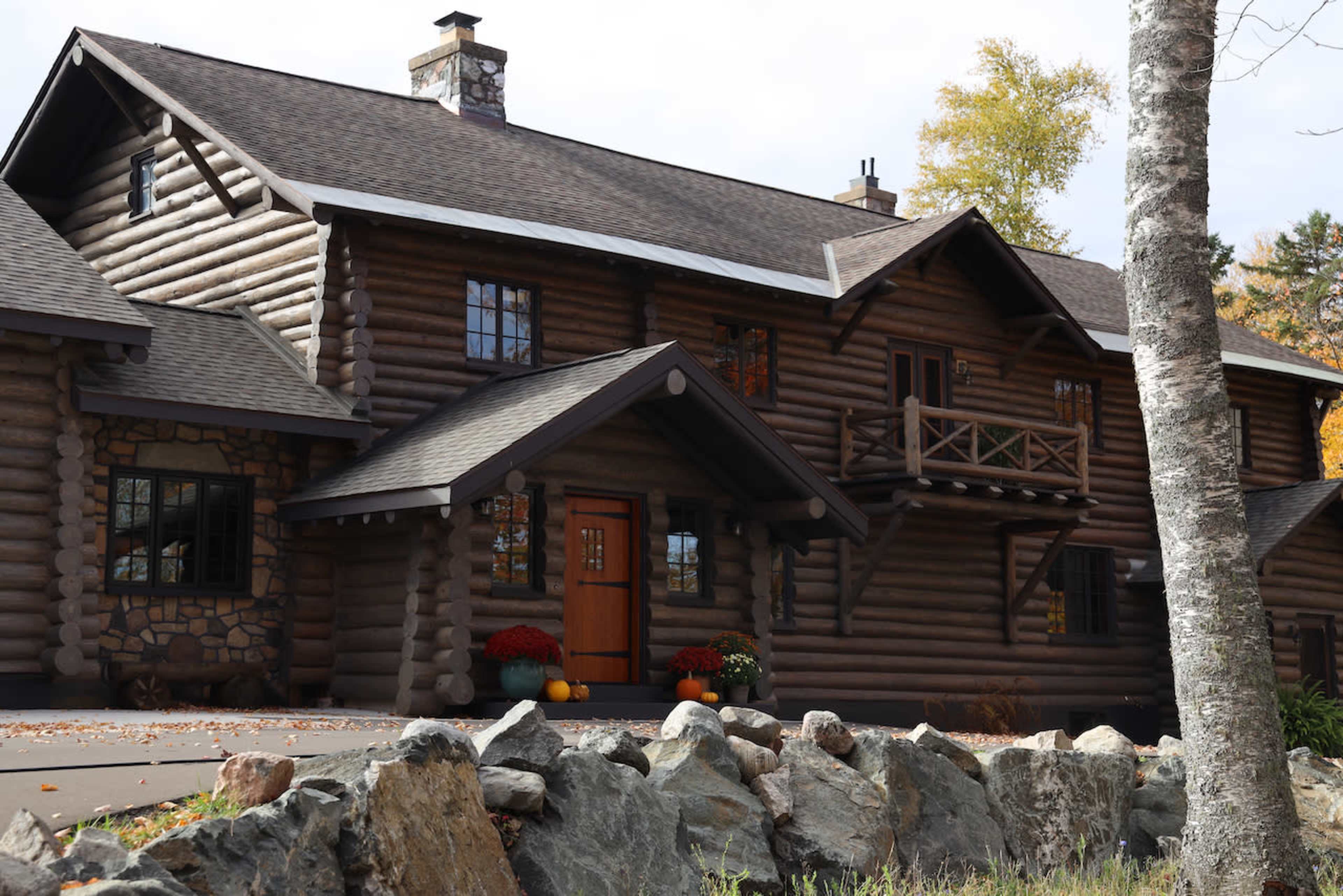 The image shows a large log cabin with a stone foundation surrounded by rocks and autumn foliage.