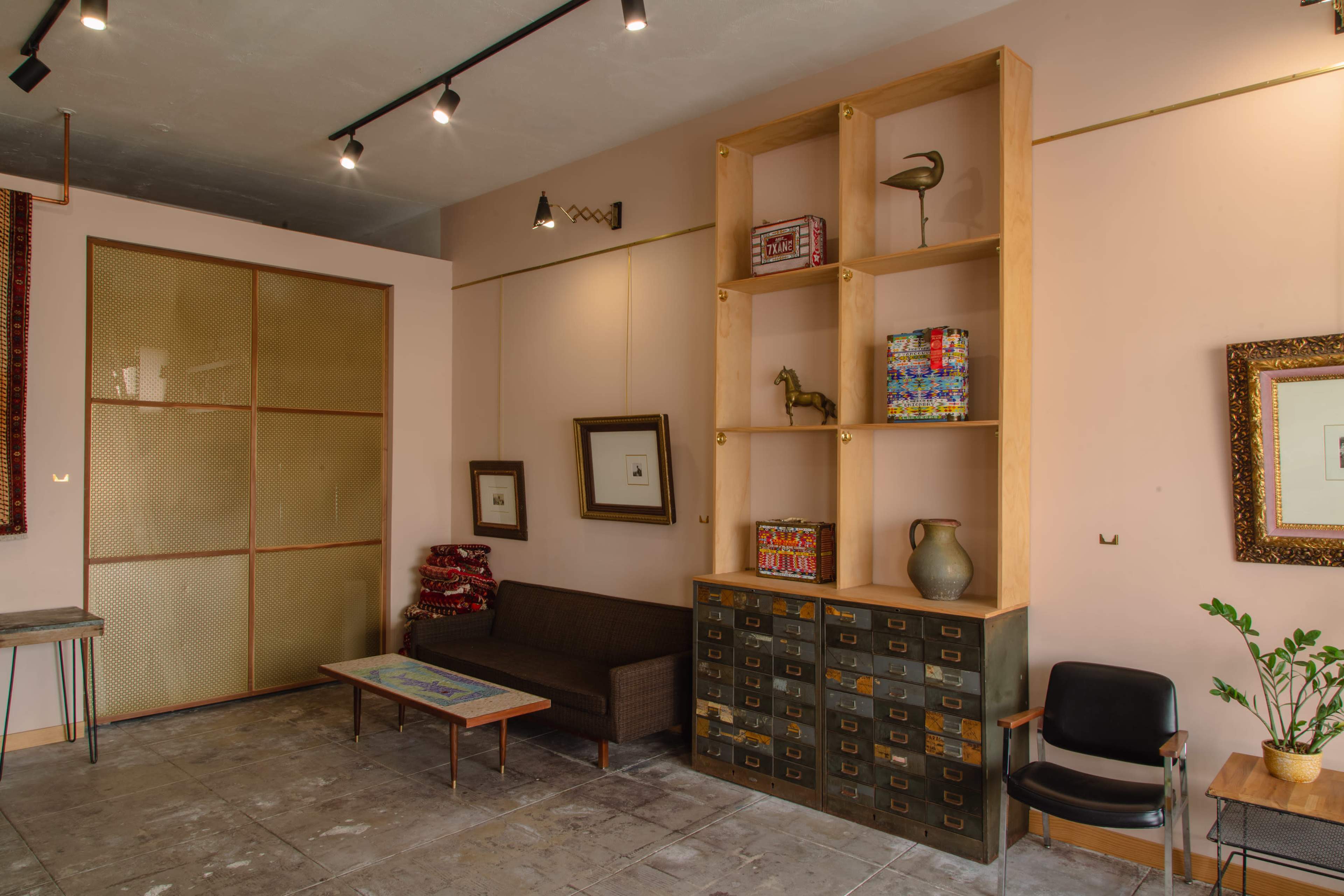 A minimalist living area features a brown sofa, a wooden shelving unit, a decorative cabinet with drawers, and a black chair against a pale wall.