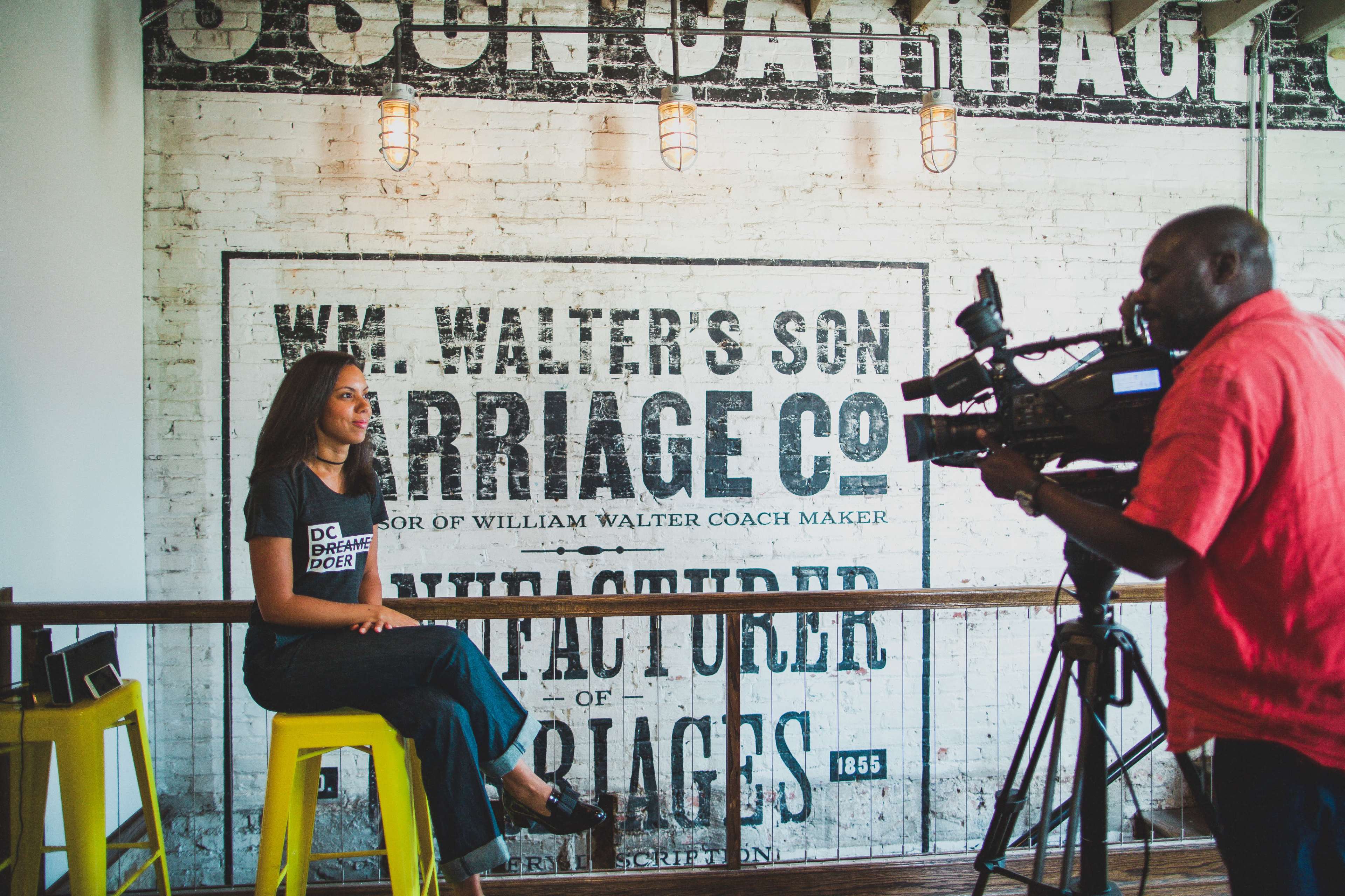 A woman sits on a yellow stool while being interviewed by a man holding a camera in front of a vintage carriage company sign.