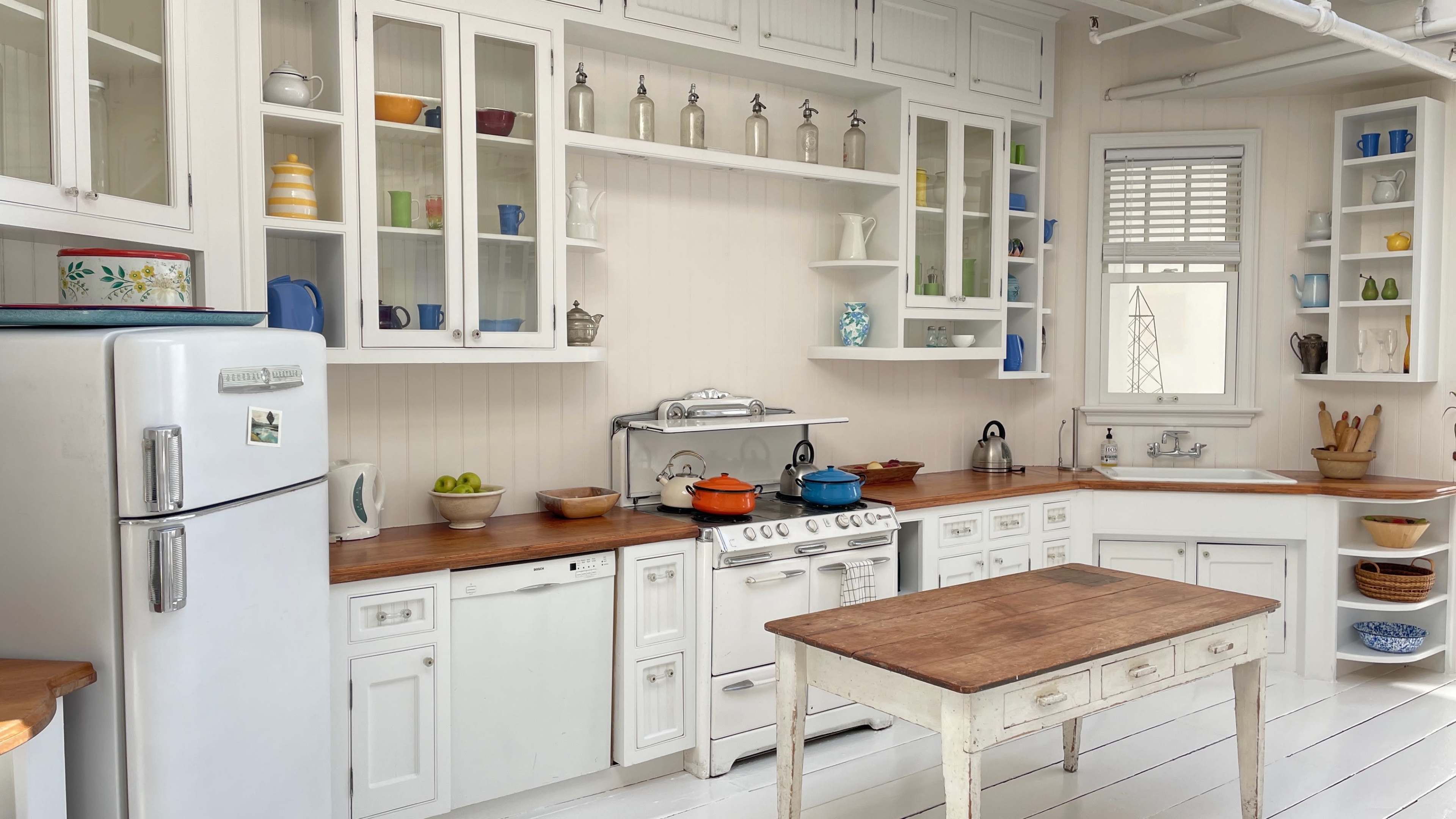 A bright, organized kitchen featuring white cabinetry, a wooden countertop, a stove, and various dishes displayed in glass-front cabinets.