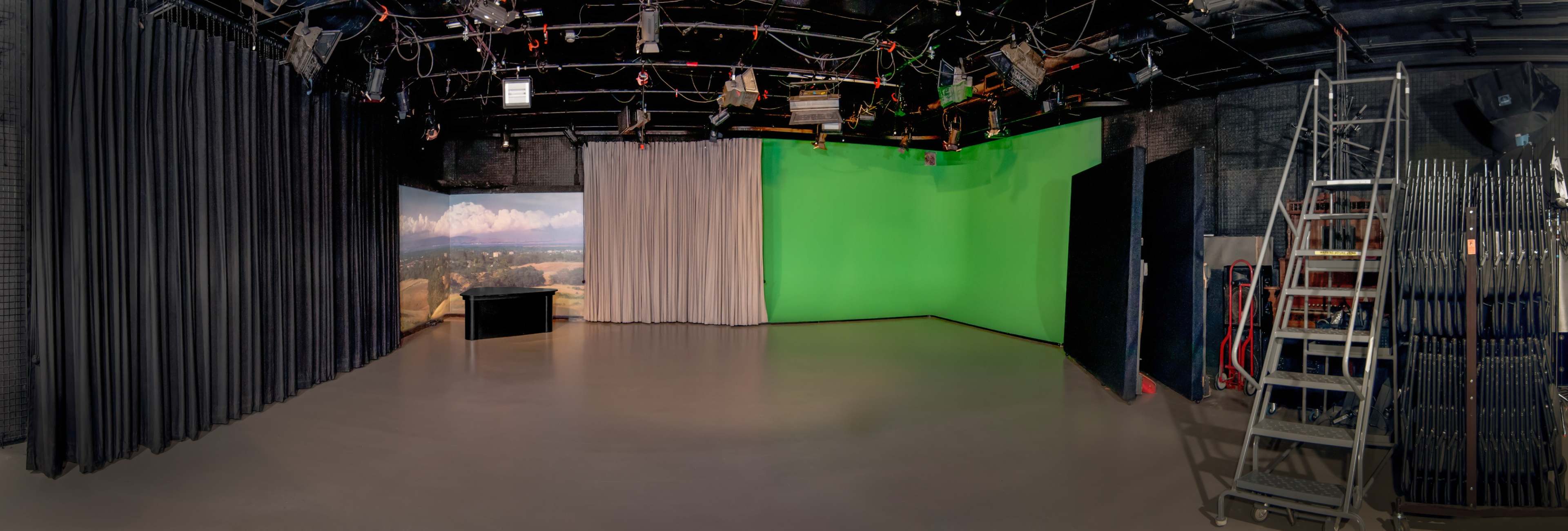 A panoramic view of a television studio with a green screen wall, a black desk, and curtains in a simple layout.