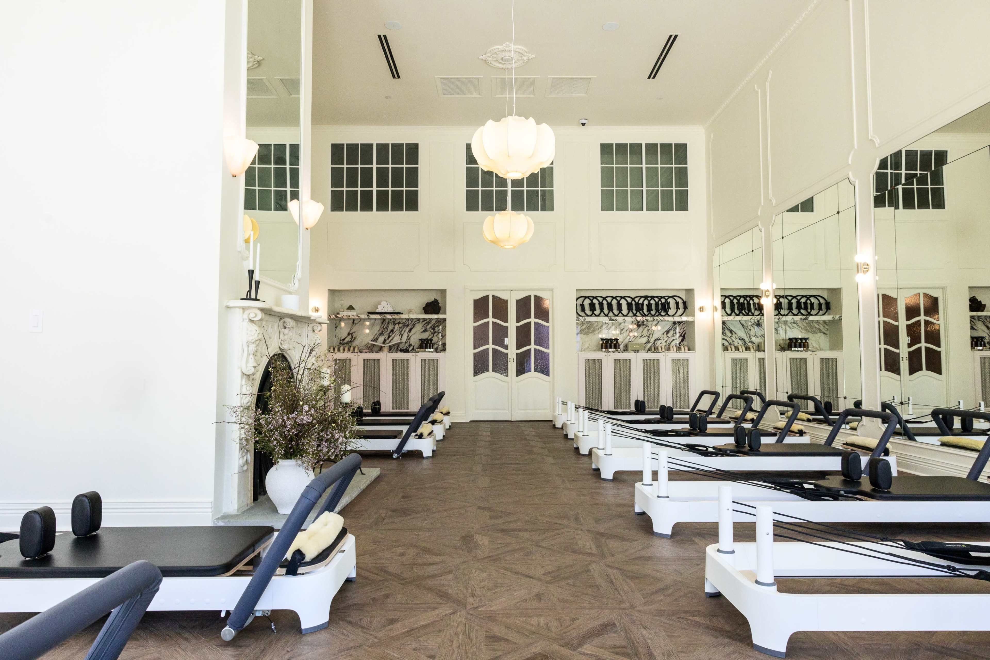 The image depicts a spacious fitness studio with several reformer machines arranged neatly in rows, featuring large windows and an elegant fireplace.