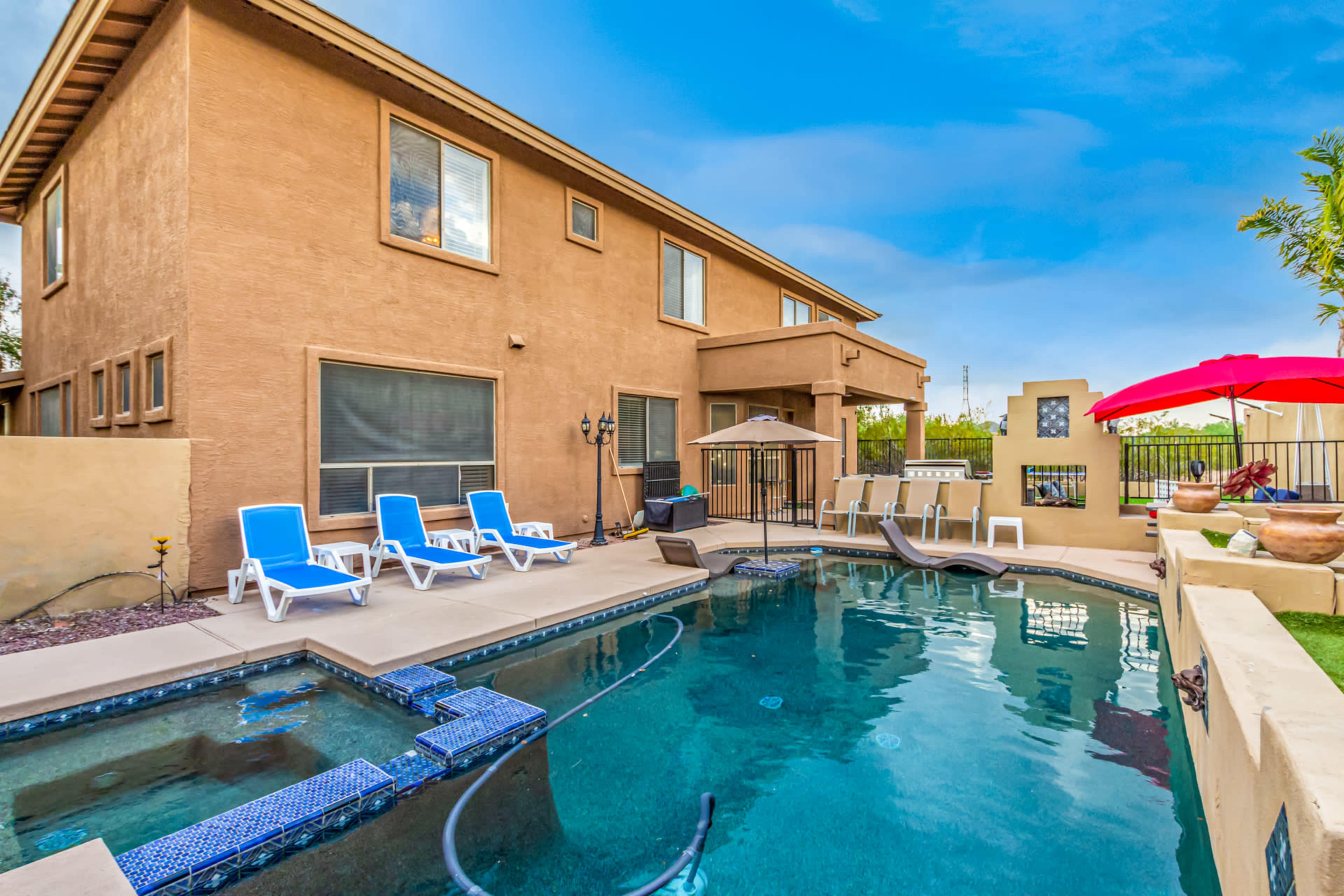 The image shows a backyard with a swimming pool surrounded by lounge chairs and a shaded area with an umbrella near a two-story house.
