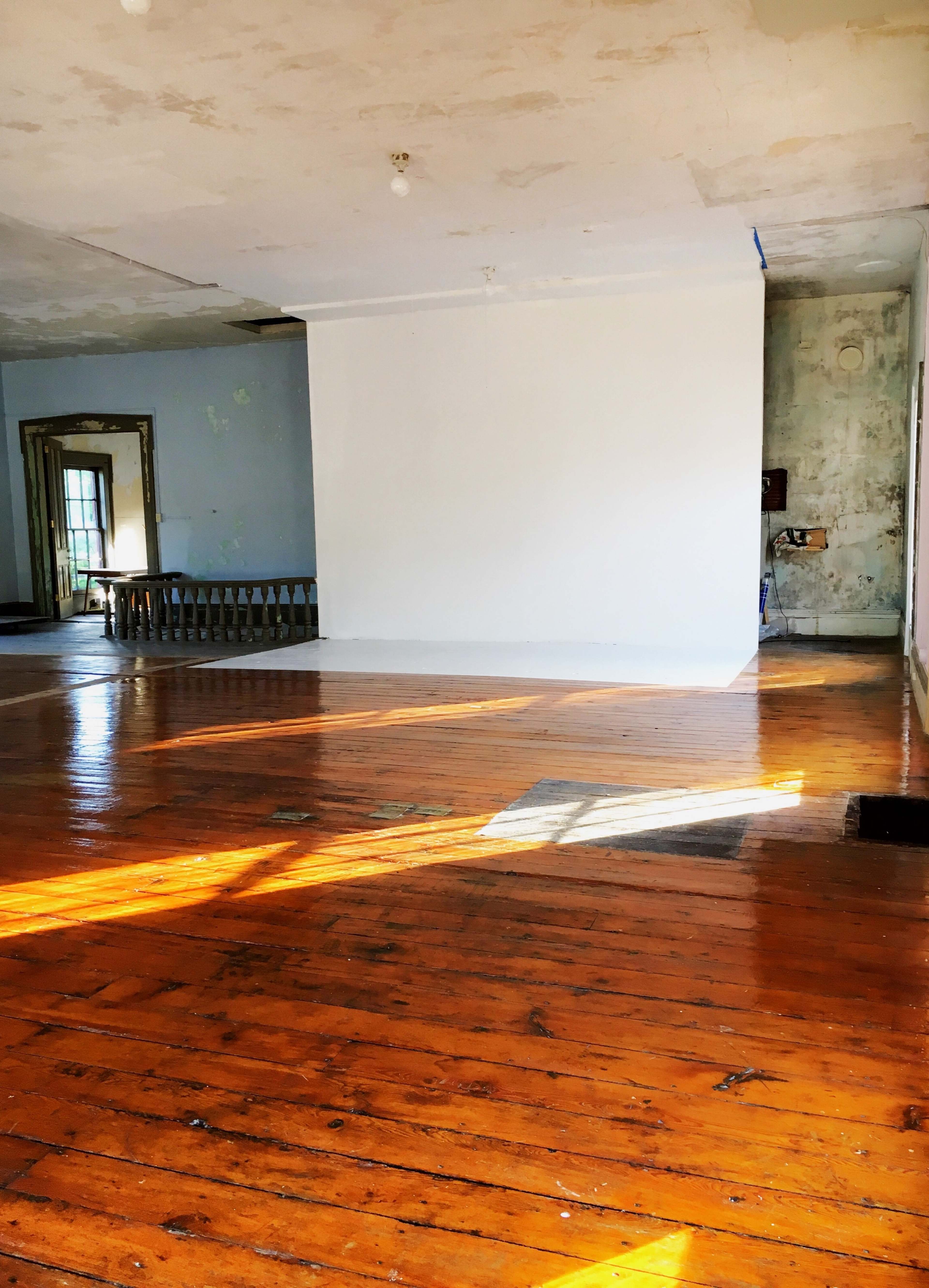 A spacious, empty room with wooden floors, a large white wall, and natural light streaming through the windows.