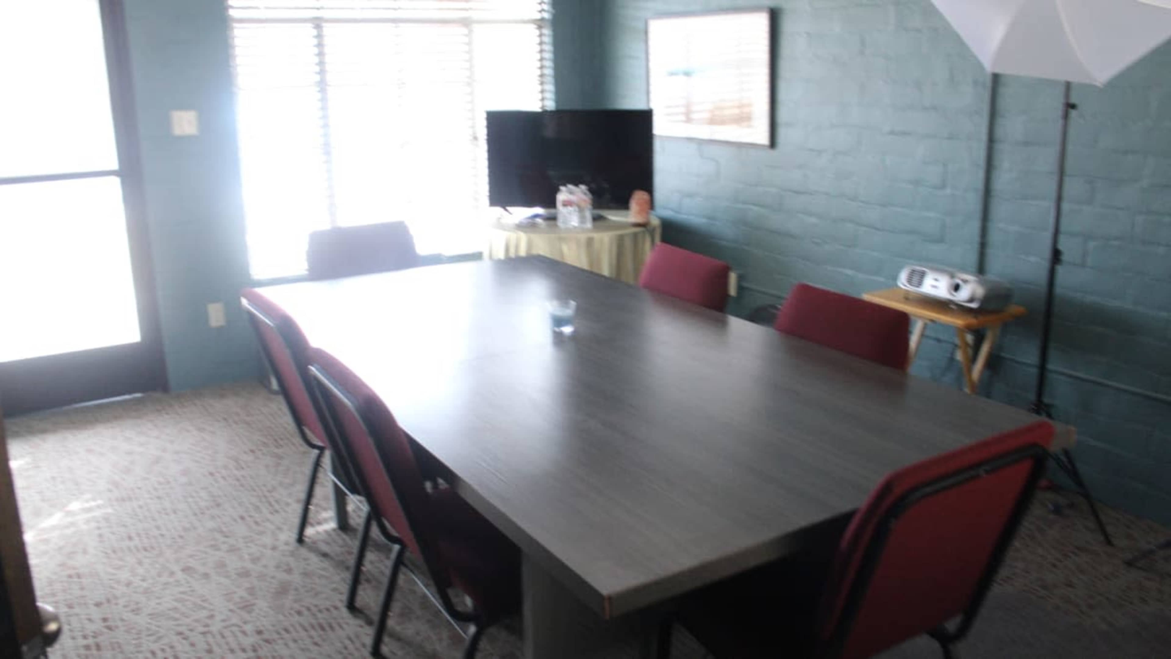 The image shows a conference room with a long table surrounded by red chairs, near a window and a television.