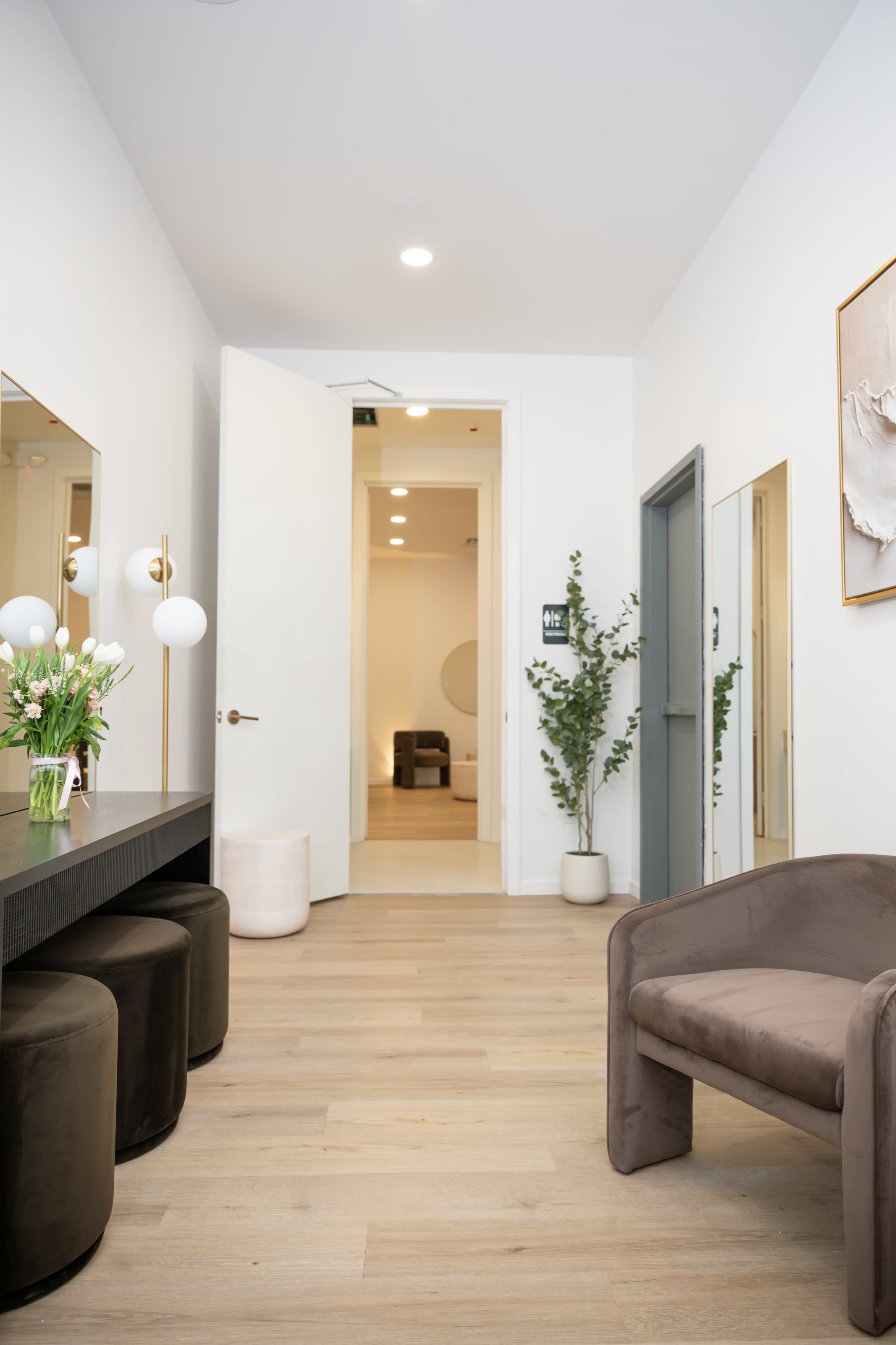 The image shows a modern, well-lit hallway leading to a room, with a mirrored vanity area on one side and a plant beside a chair on the other.