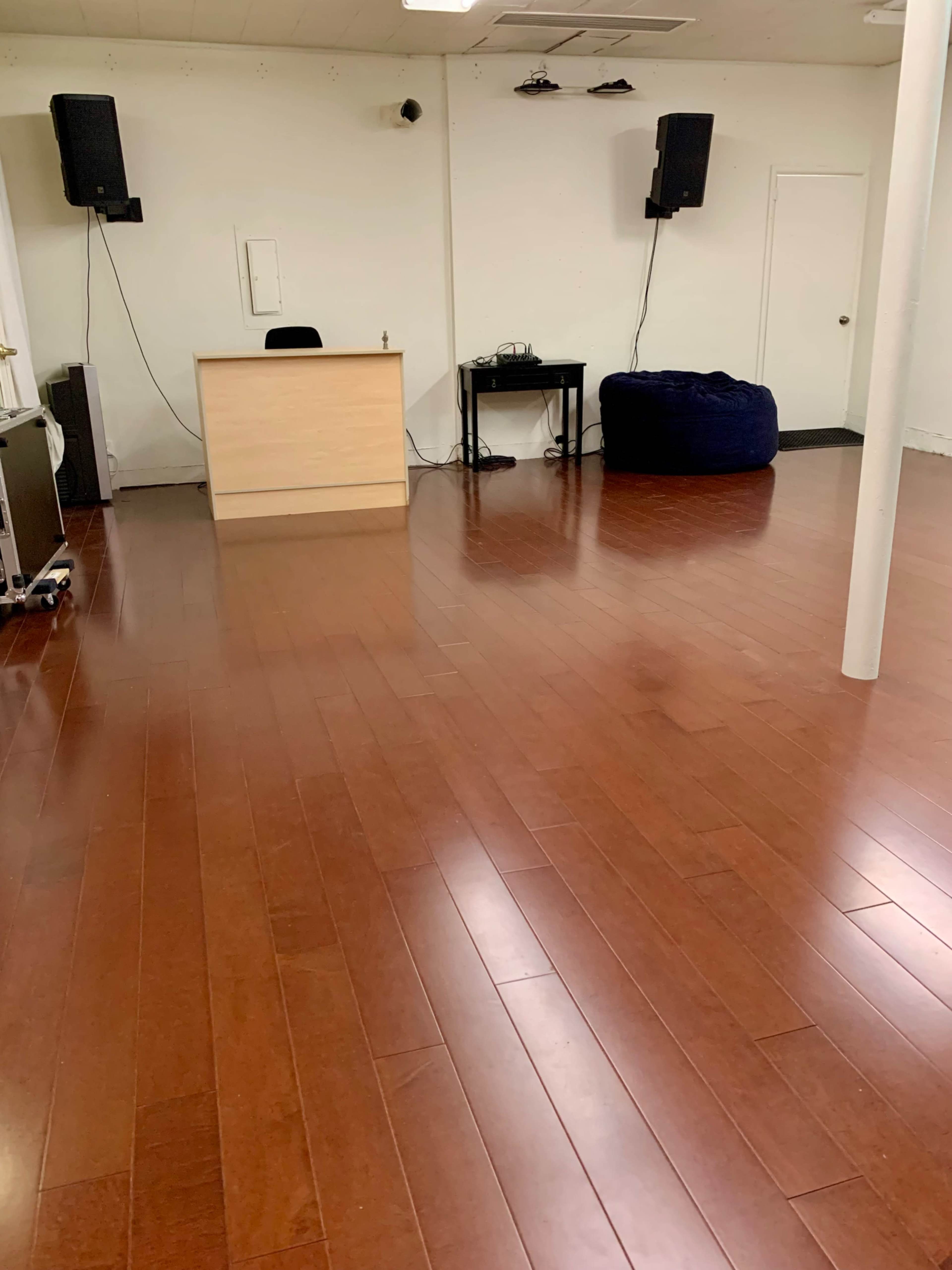 The image shows an empty room with wooden flooring, featuring a reception desk, a small black table with equipment, and a blue bean bag in the corner.