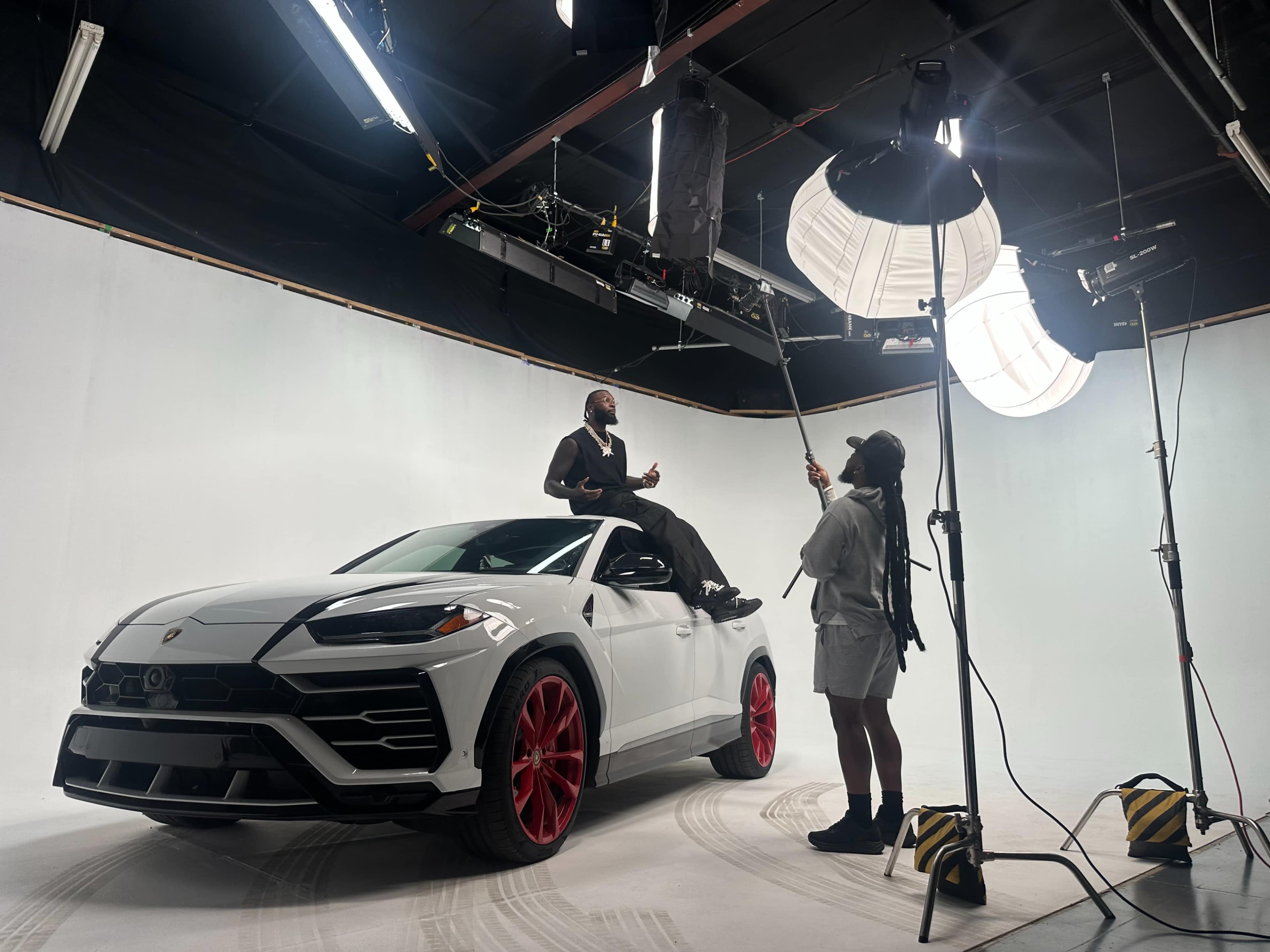 A man is sitting on top of a white luxury SUV in a photography studio, while a woman adjusts lighting equipment.