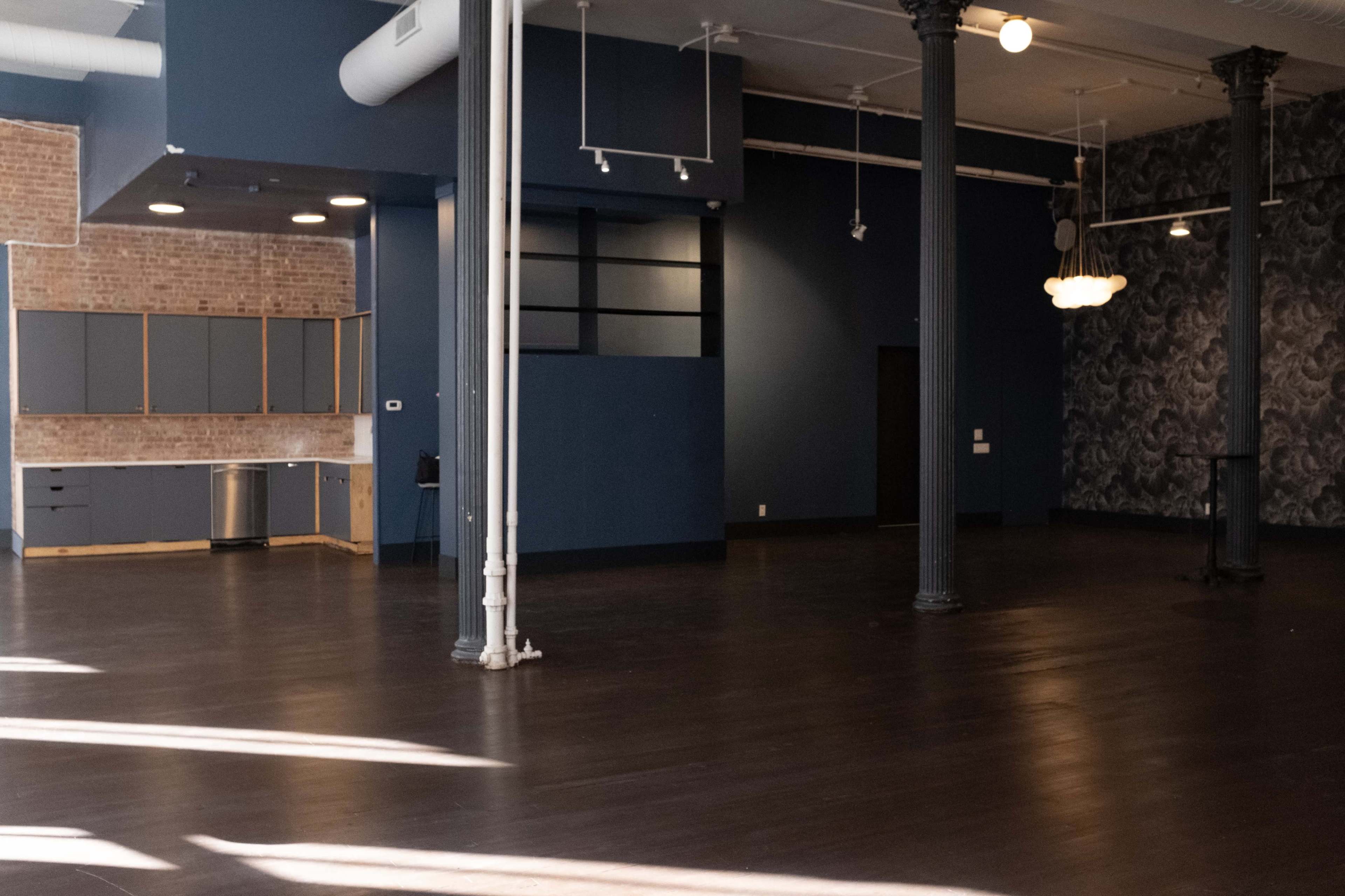 The image shows a spacious, empty room with hardwood floors, tall ceilings, and a combination of exposed brick and dark blue walls.