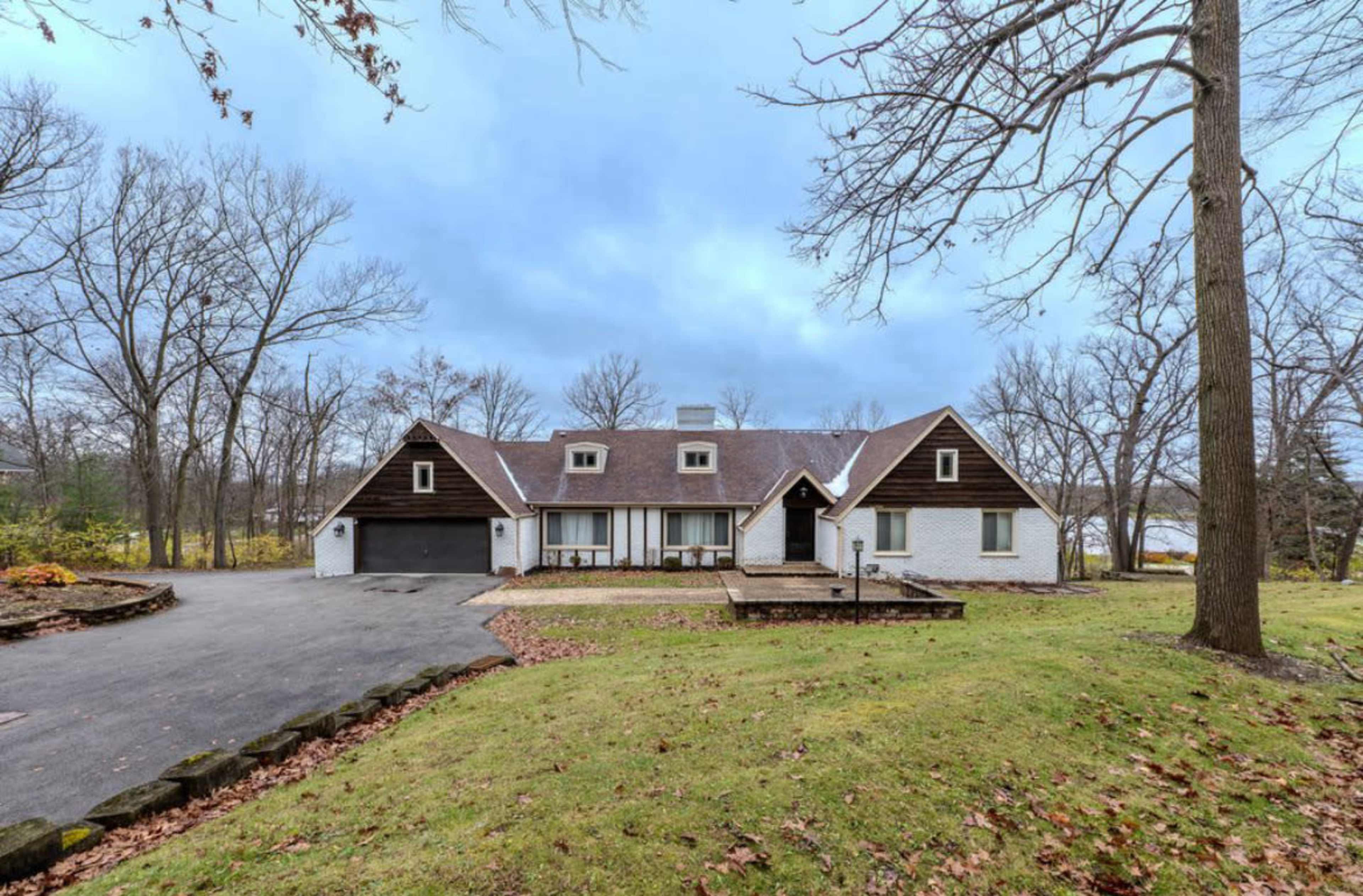 A large, two-story home with a symmetrical roofline is set in a wooded area, featuring a paved driveway and a front porch.