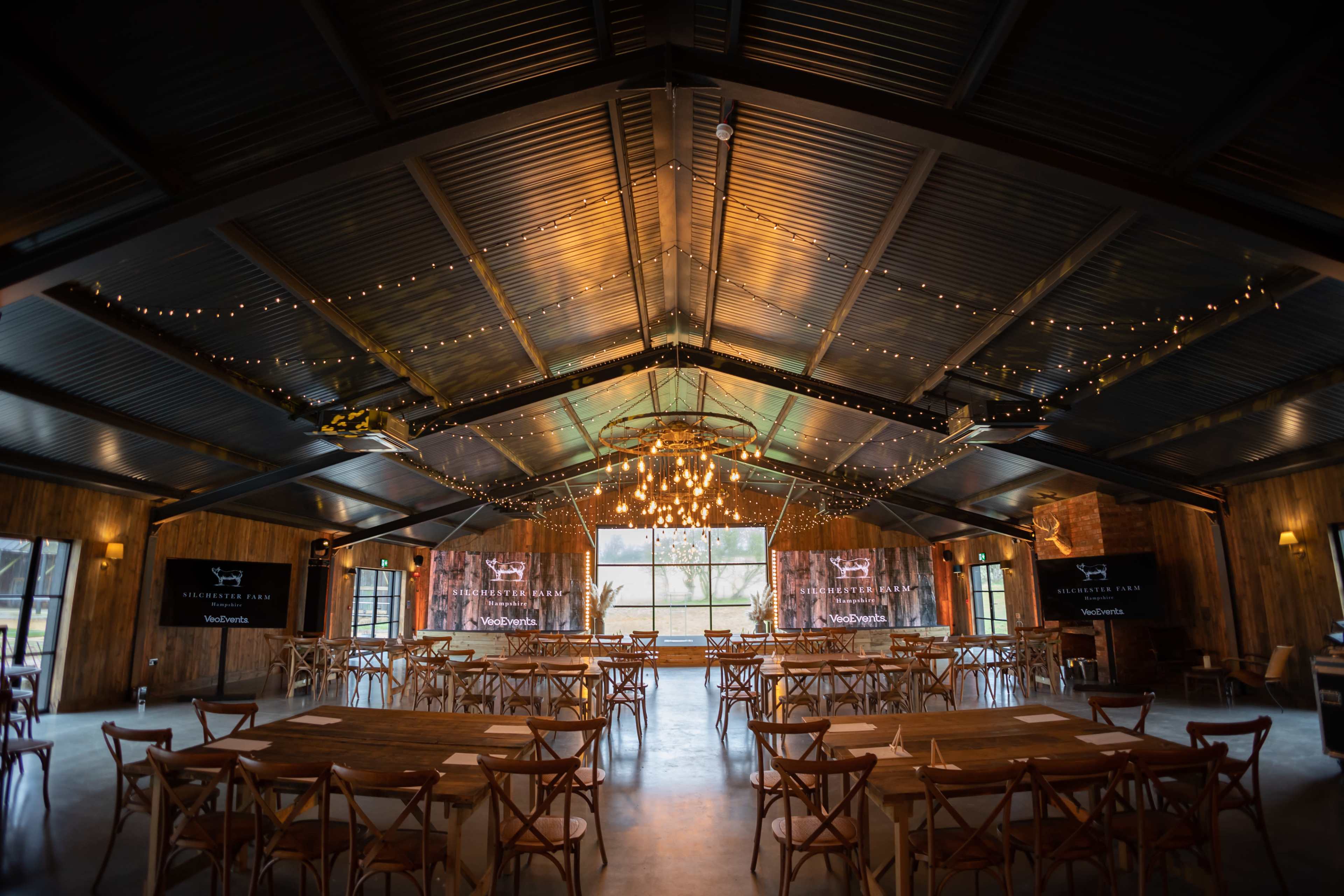 The interior of a spacious event venue features wooden tables arranged in rows, with string lights and a large chandelier illuminating the ceiling.