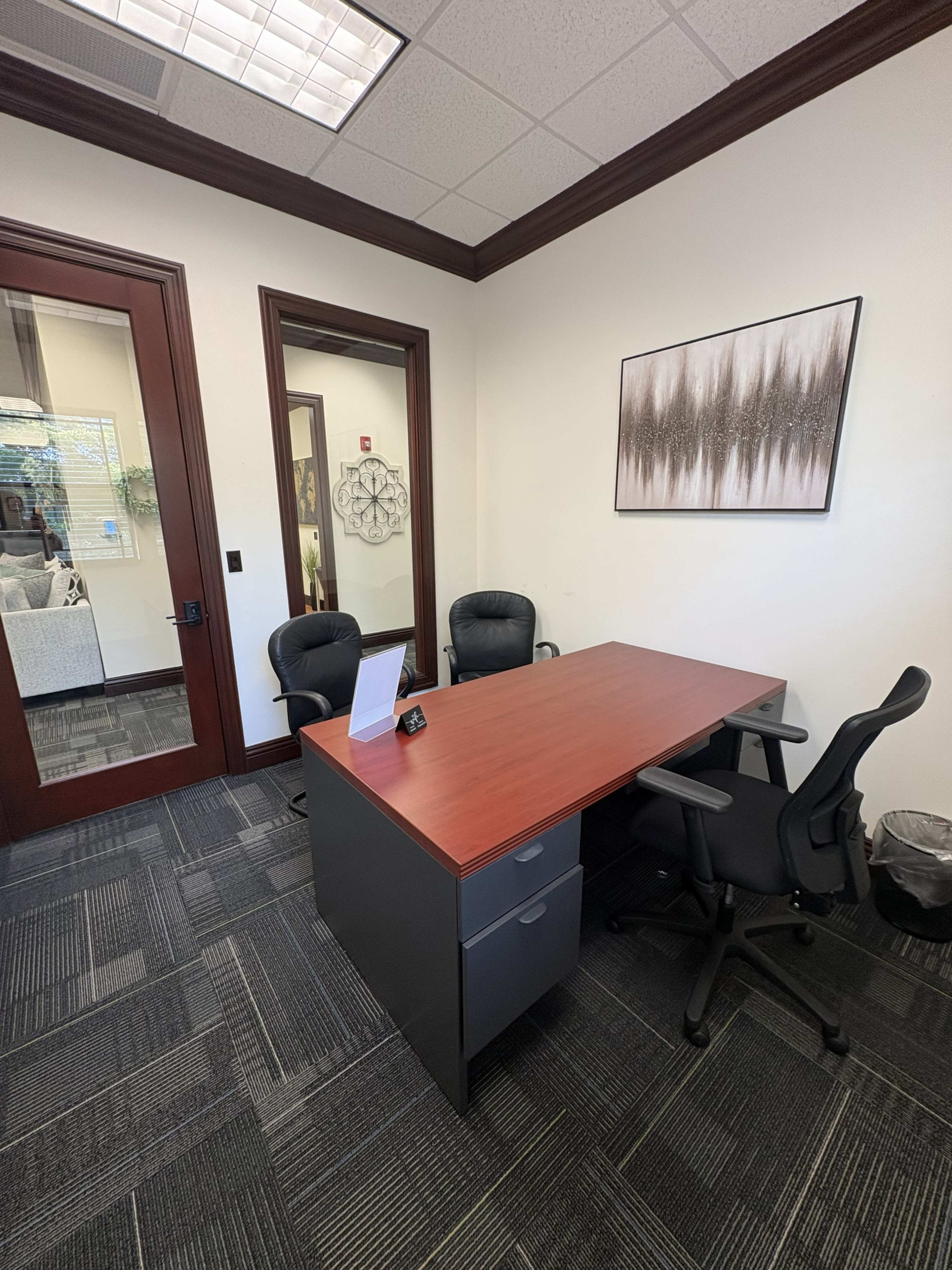 The image shows a small office space with a wooden desk, two black office chairs, a framed artwork on the wall, and a partial view of another room through a glass door.