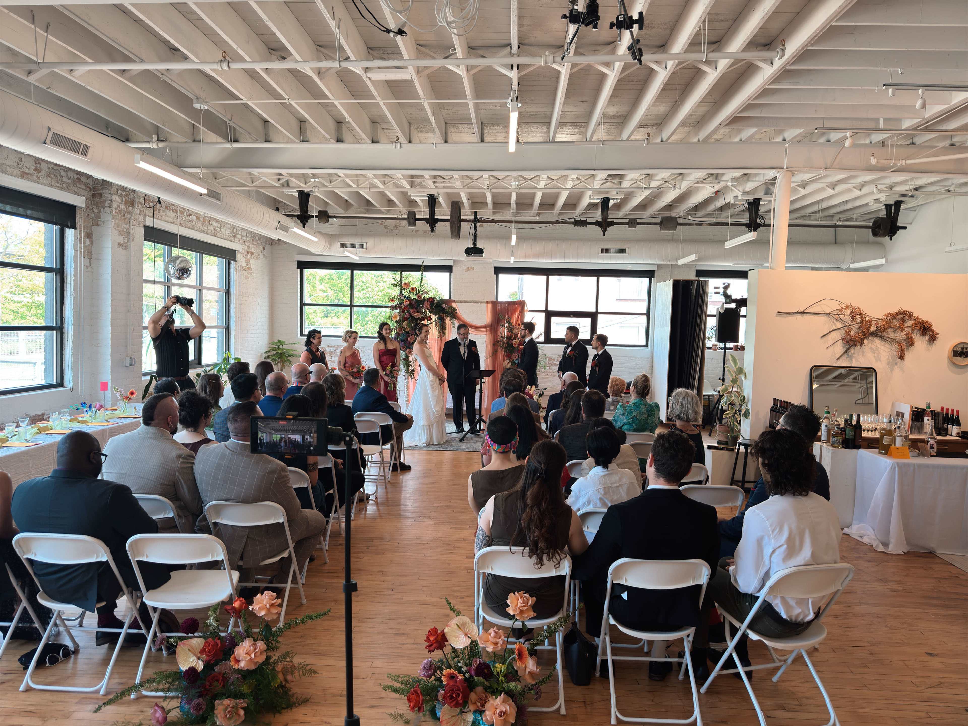 A wedding ceremony takes place in a bright, modern venue with guests seated in rows and a couple standing at the front, flanked by a decorated backdrop.