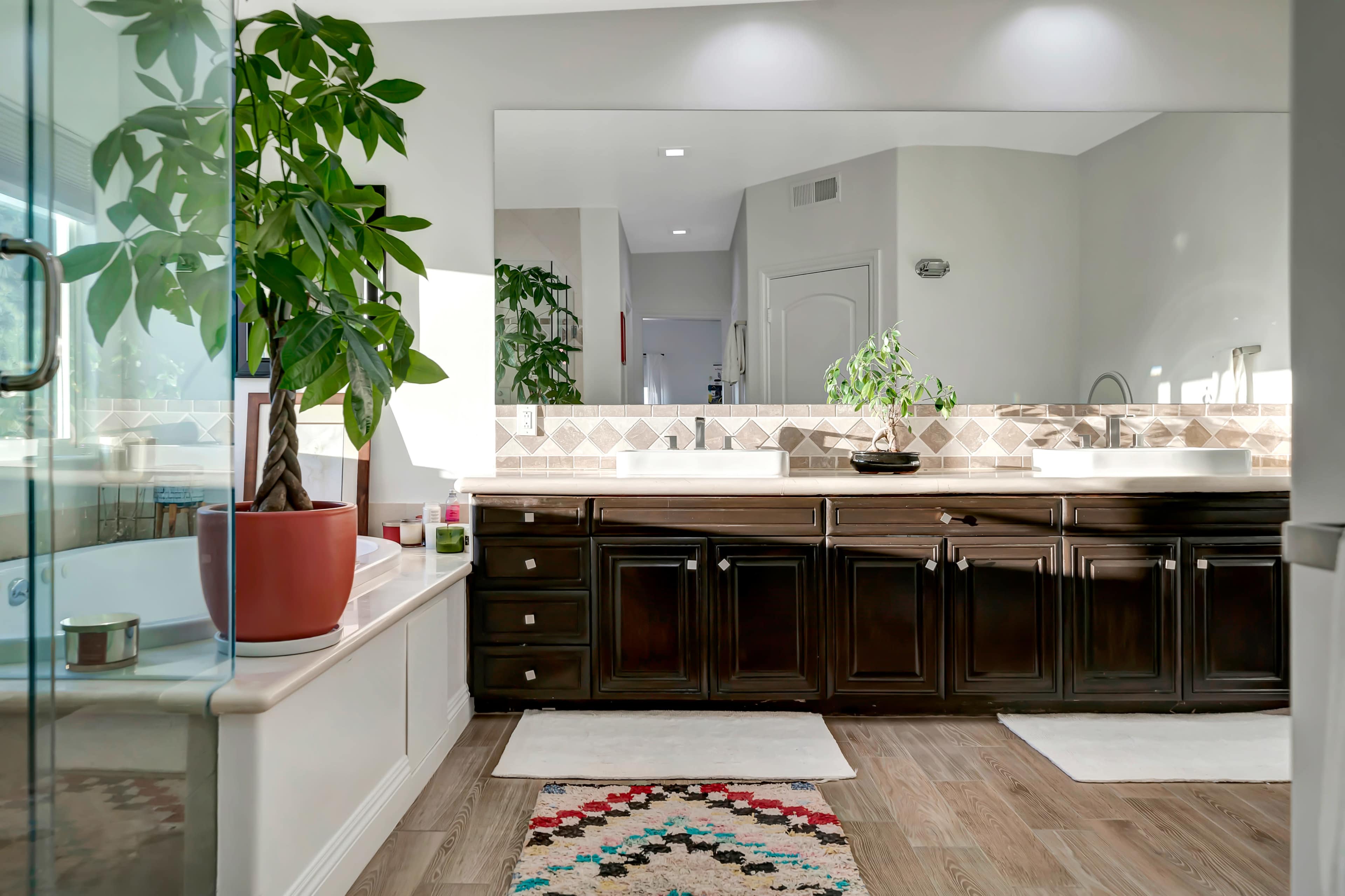 The image depicts a modern bathroom featuring a large double vanity with dark wood cabinetry, a soaking tub, and a green plant positioned near a window.