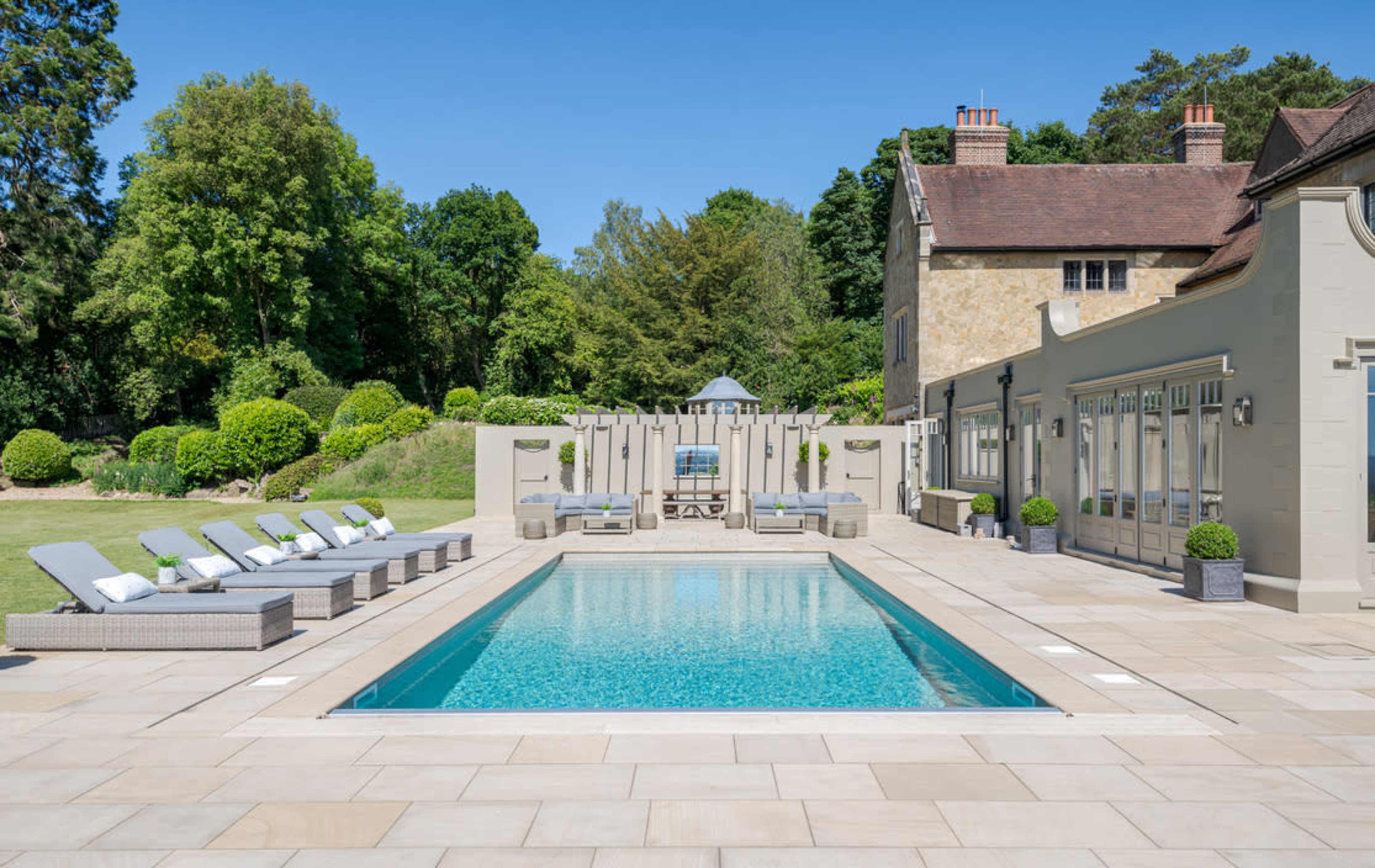 The image shows a rectangular swimming pool surrounded by lounge chairs and a garden with lush greenery and a large house in the background.