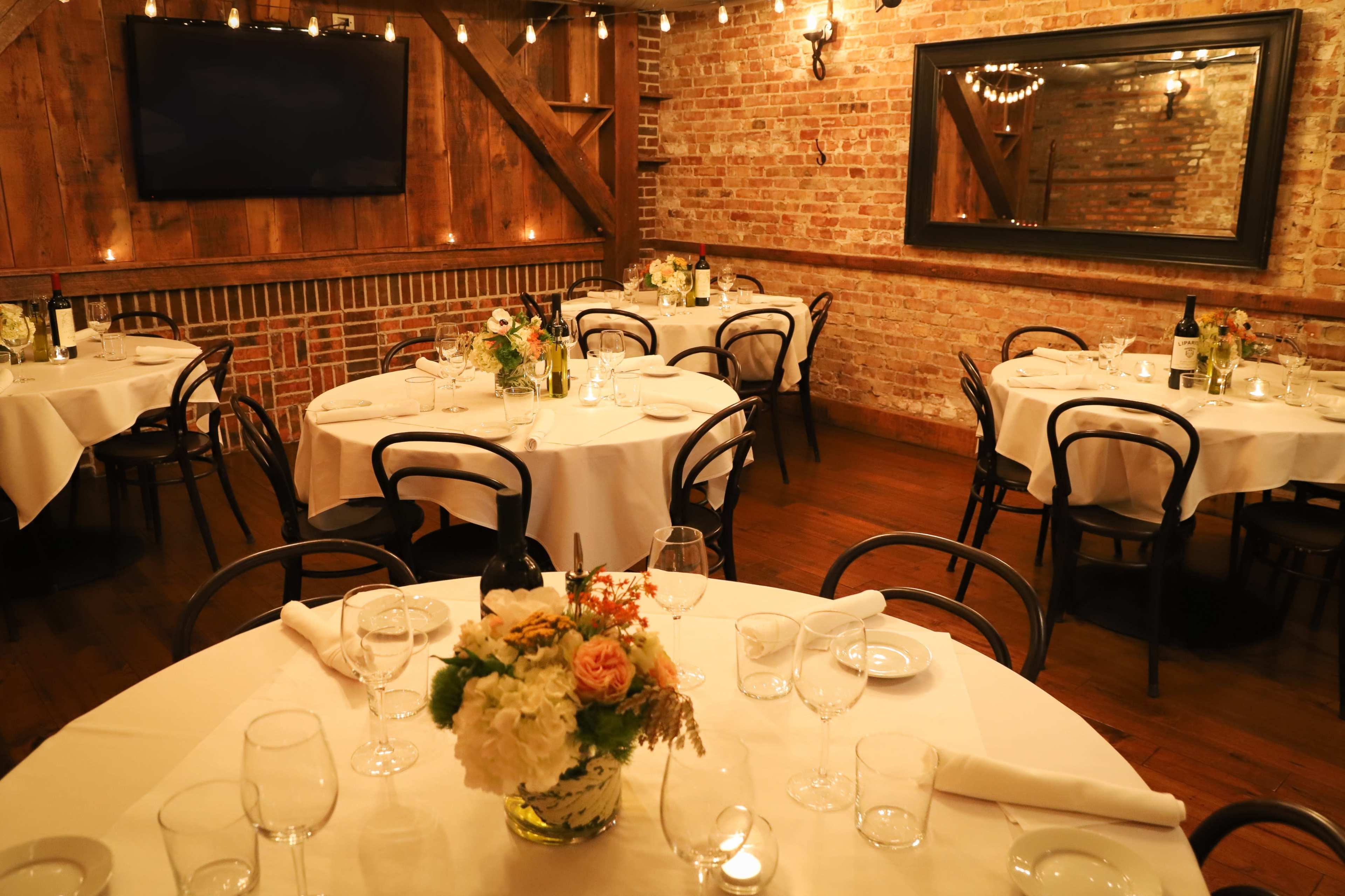 The image shows a cozy restaurant interior with round tables set for dining, featuring floral centerpieces and soft lighting.