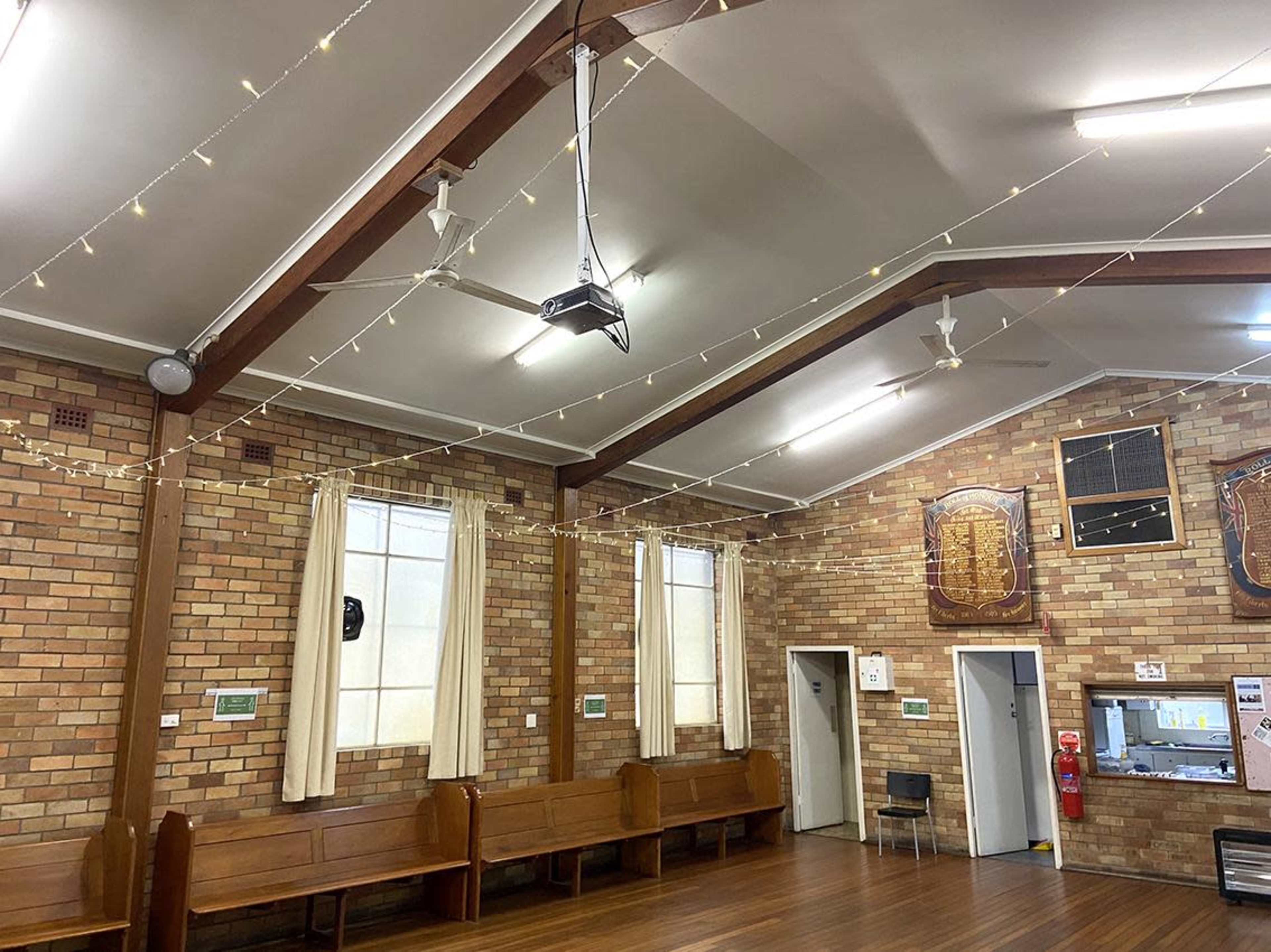 The image shows the interior of a hall with wooden benches, high ceilings, and string lights, featuring brick walls and a small kitchenette area.