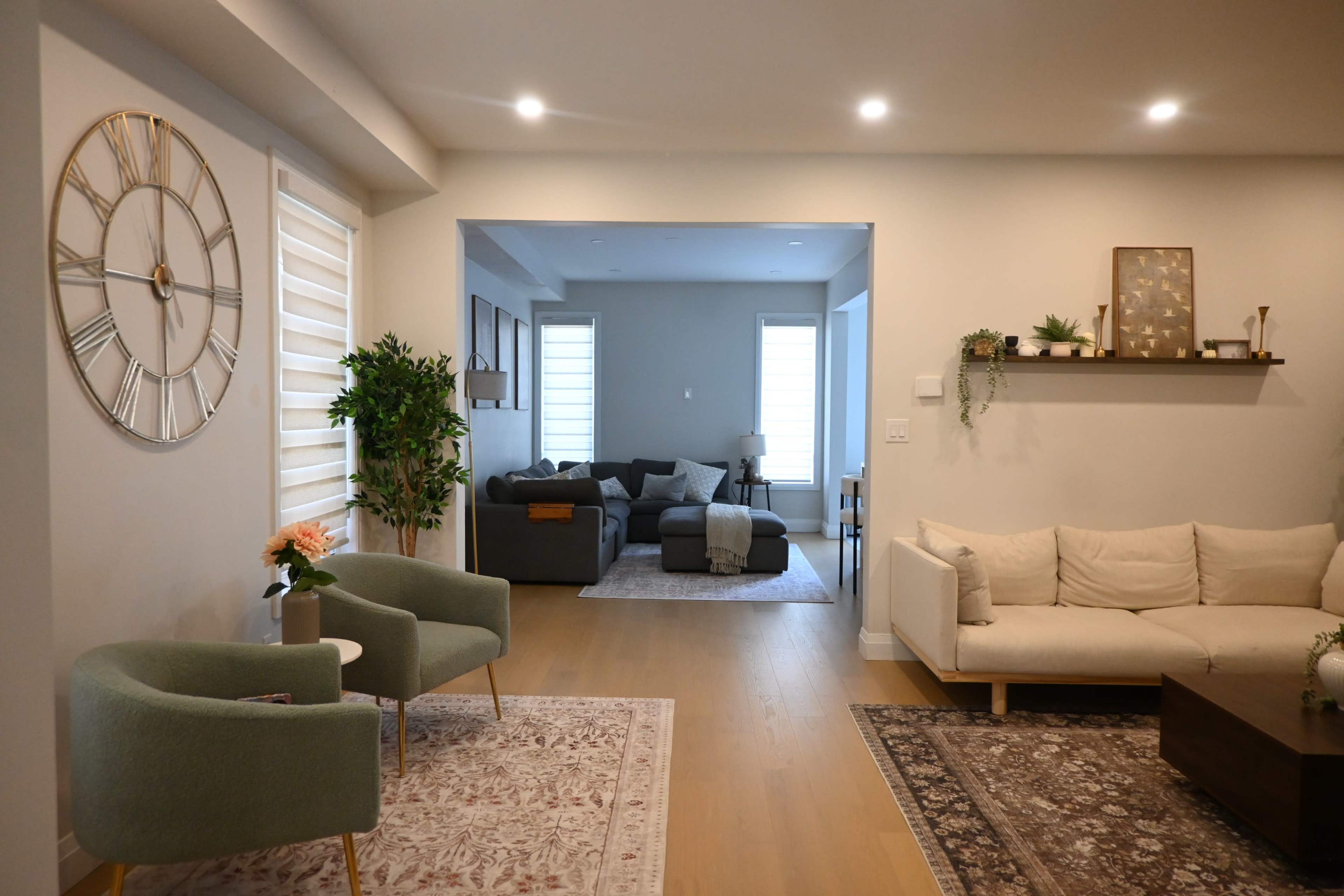 The image shows a modern living space featuring a cozy seating area with two green chairs and a large clock on the wall, leading into a separate room with a gray couch and a coffee table.