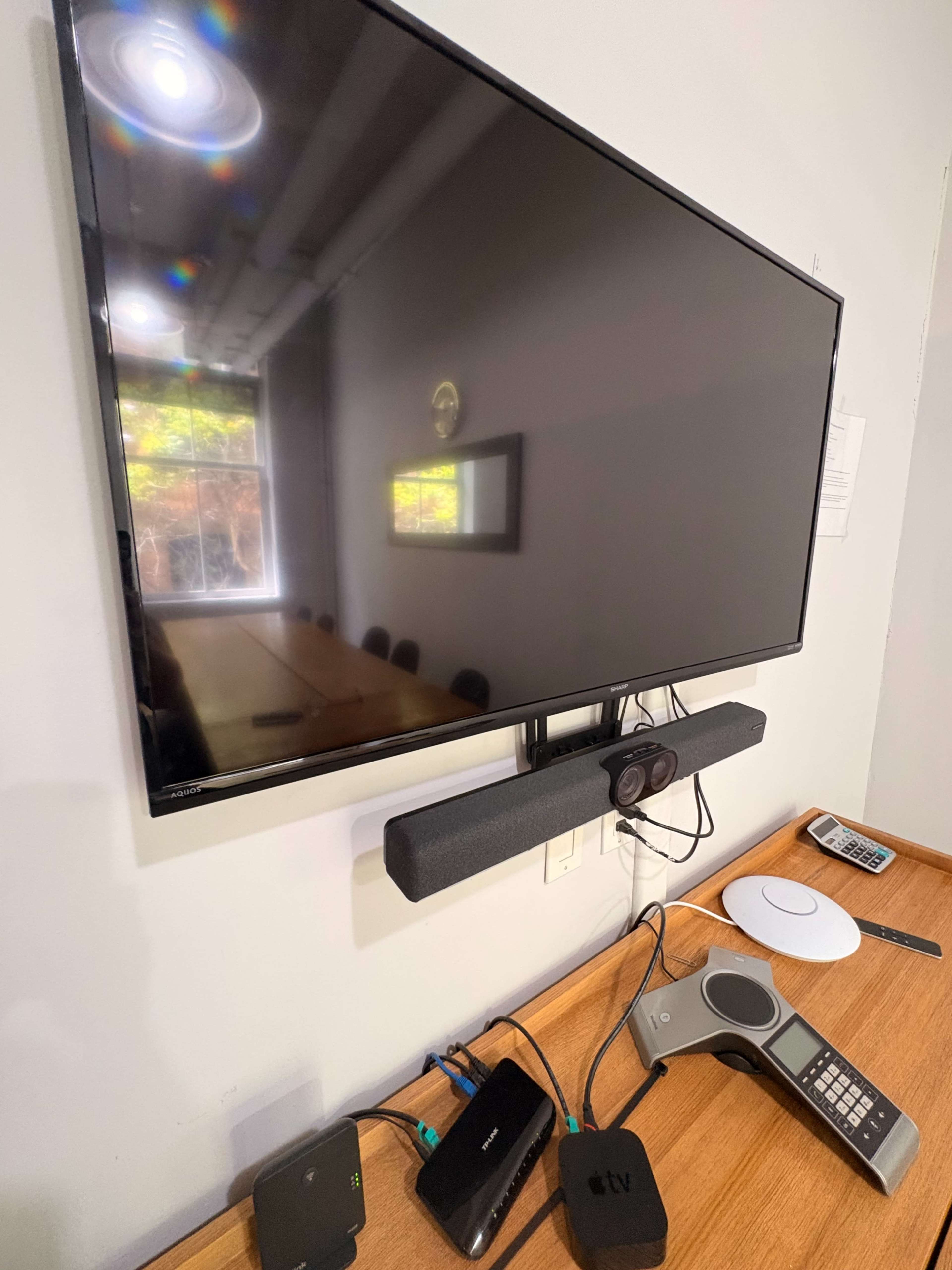 The image shows a mounted television in a conference room with a soundbar, a video conferencing device, and various electronic connections on a wooden table.