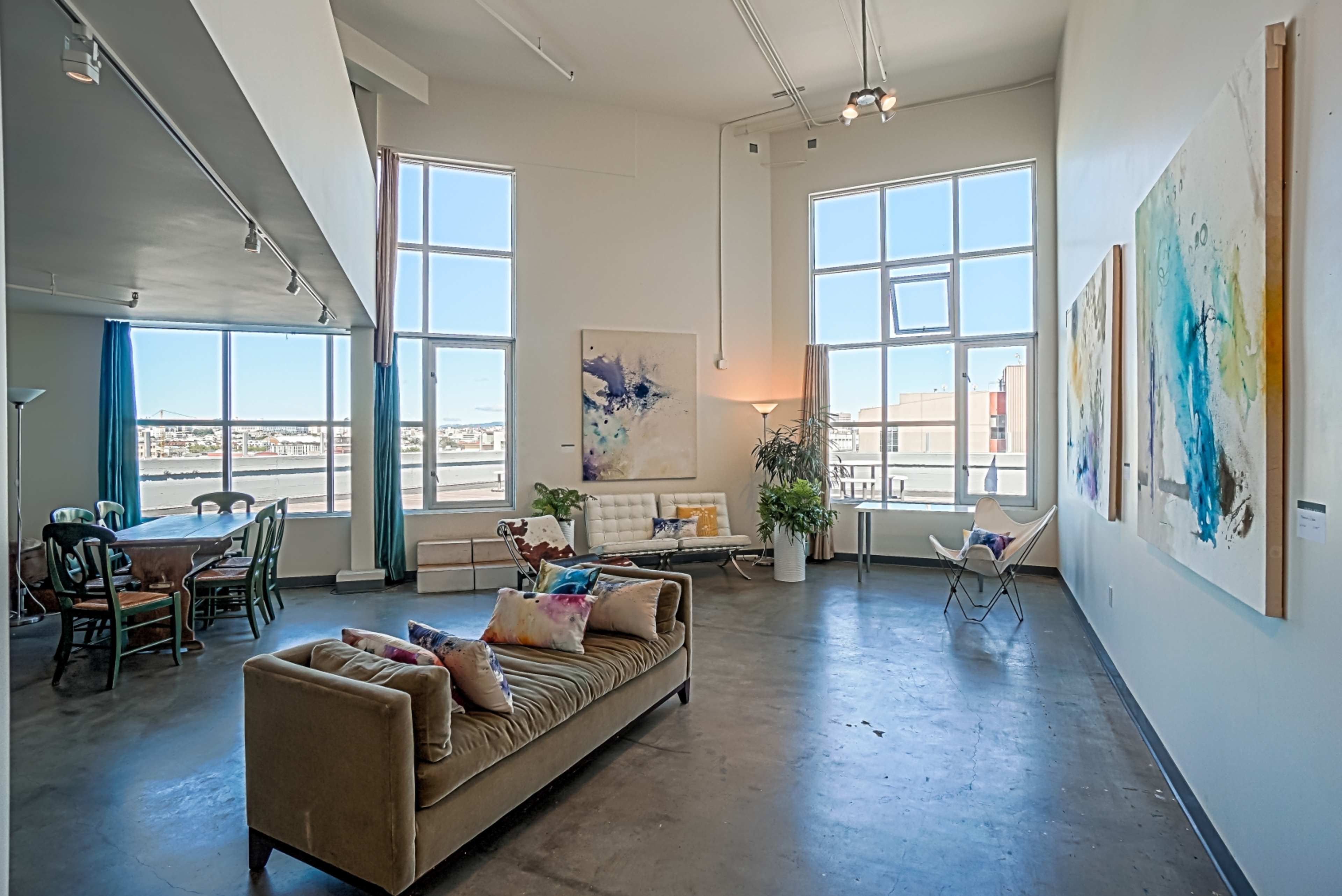 A spacious living area features large windows, a sofa with colorful pillows, a dining table, and abstract artwork on the walls.