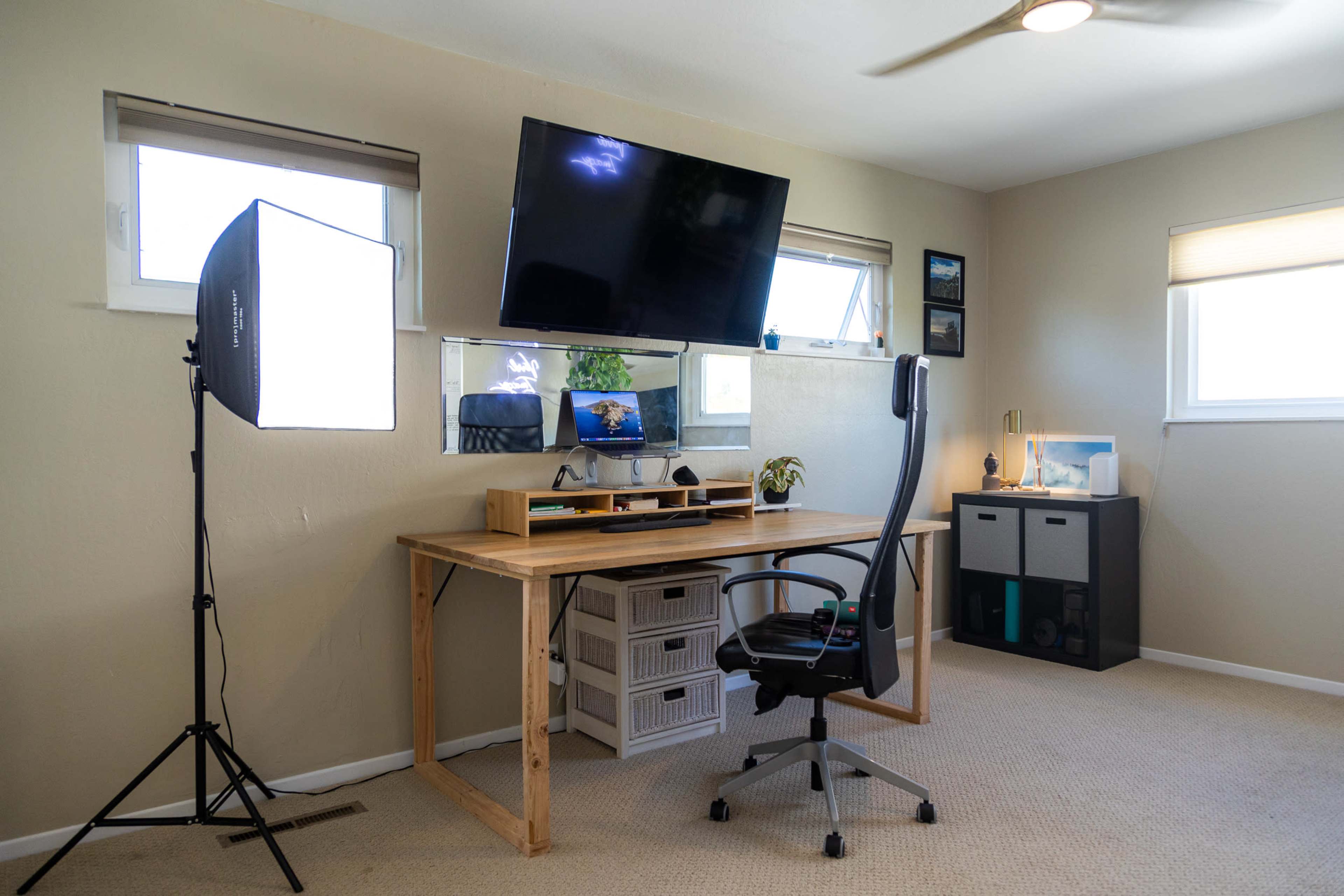 A minimalist home office setup featuring a wooden desk, an ergonomic chair, a wall-mounted television, and a studio light stand.