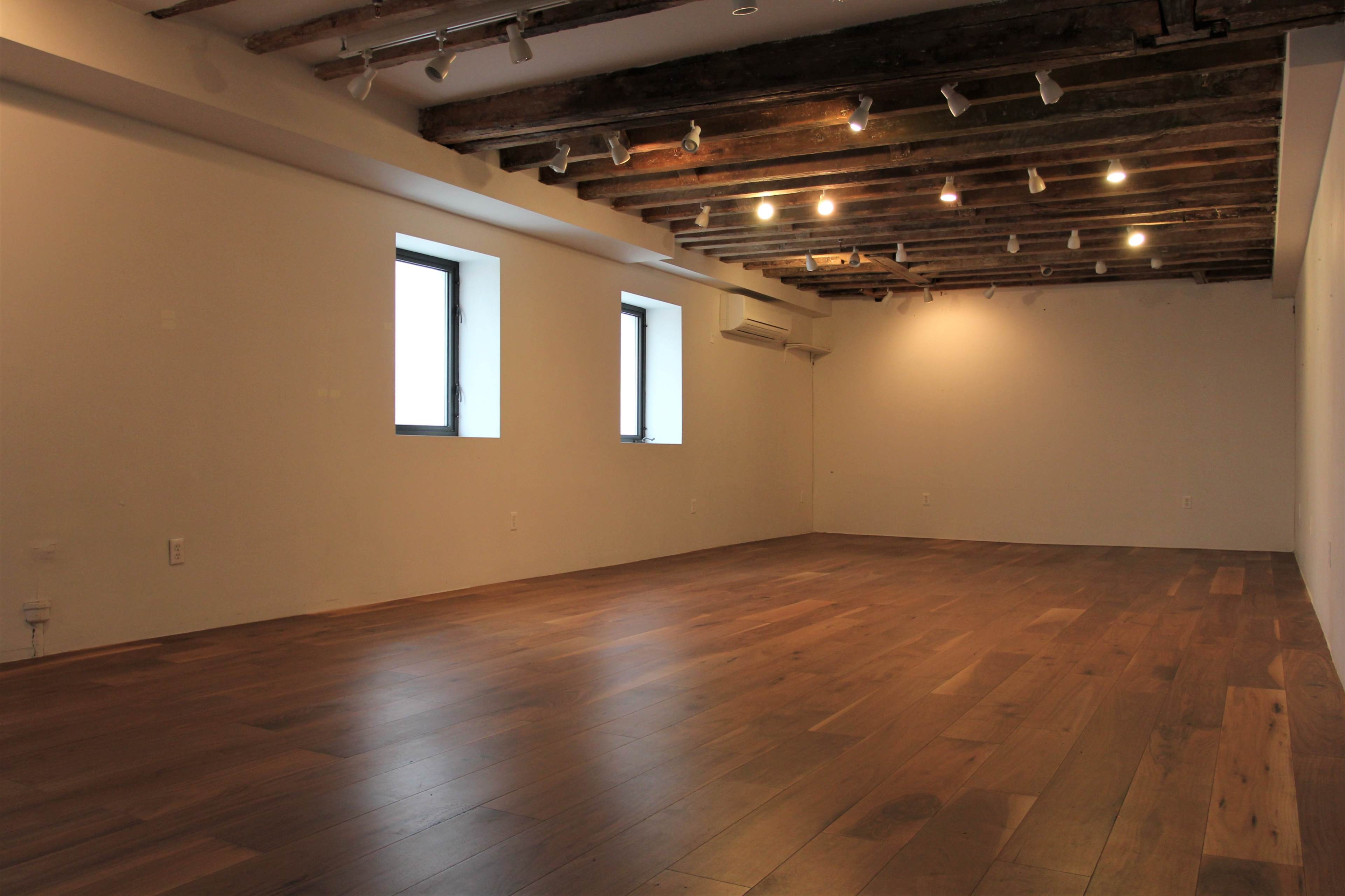 An empty room with wooden floors and exposed ceiling beams, featuring two windows and track lighting.