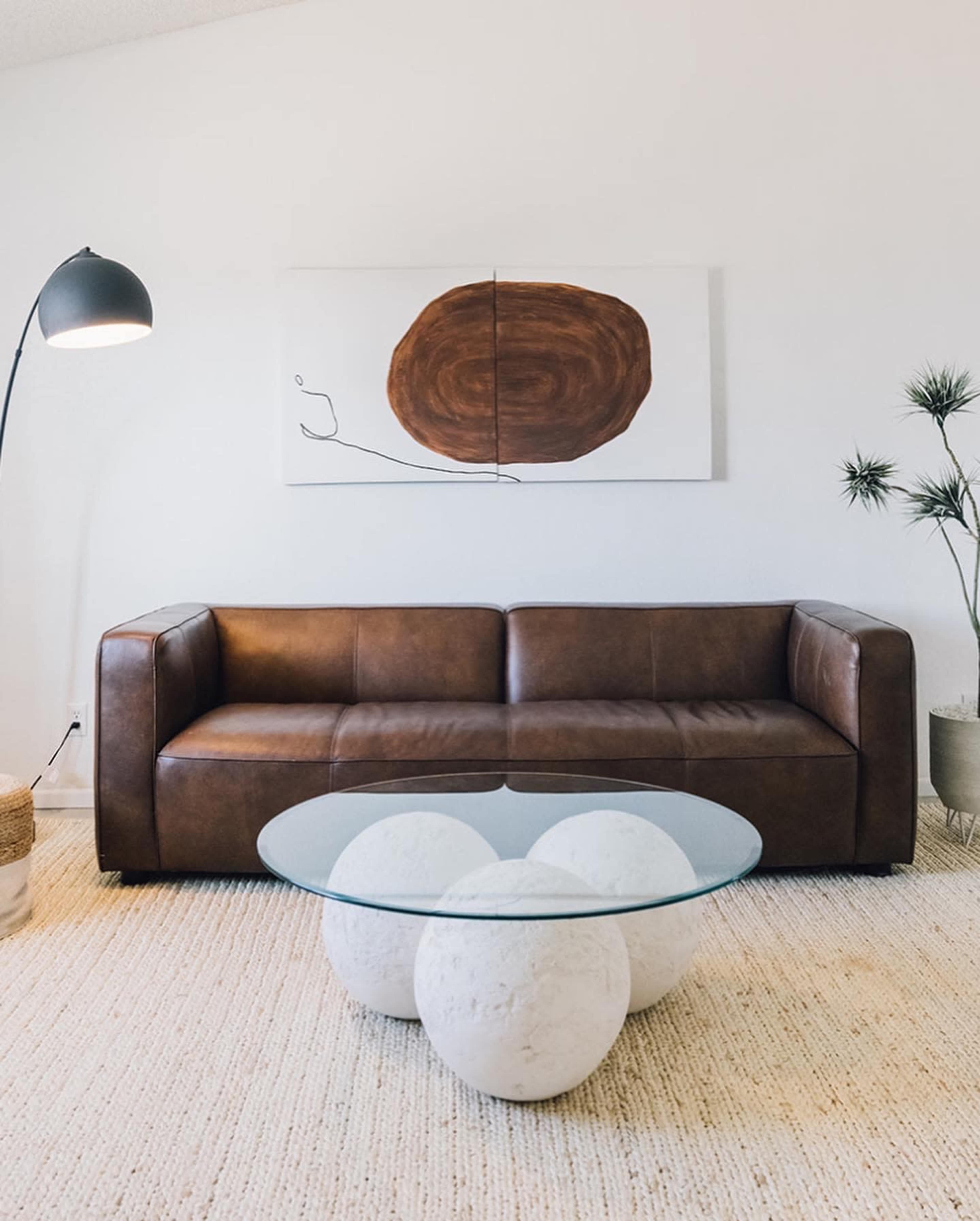 A brown leather sofa paired with a circular glass coffee table, set against a minimalist light-colored wall and adorned with abstract wall art.