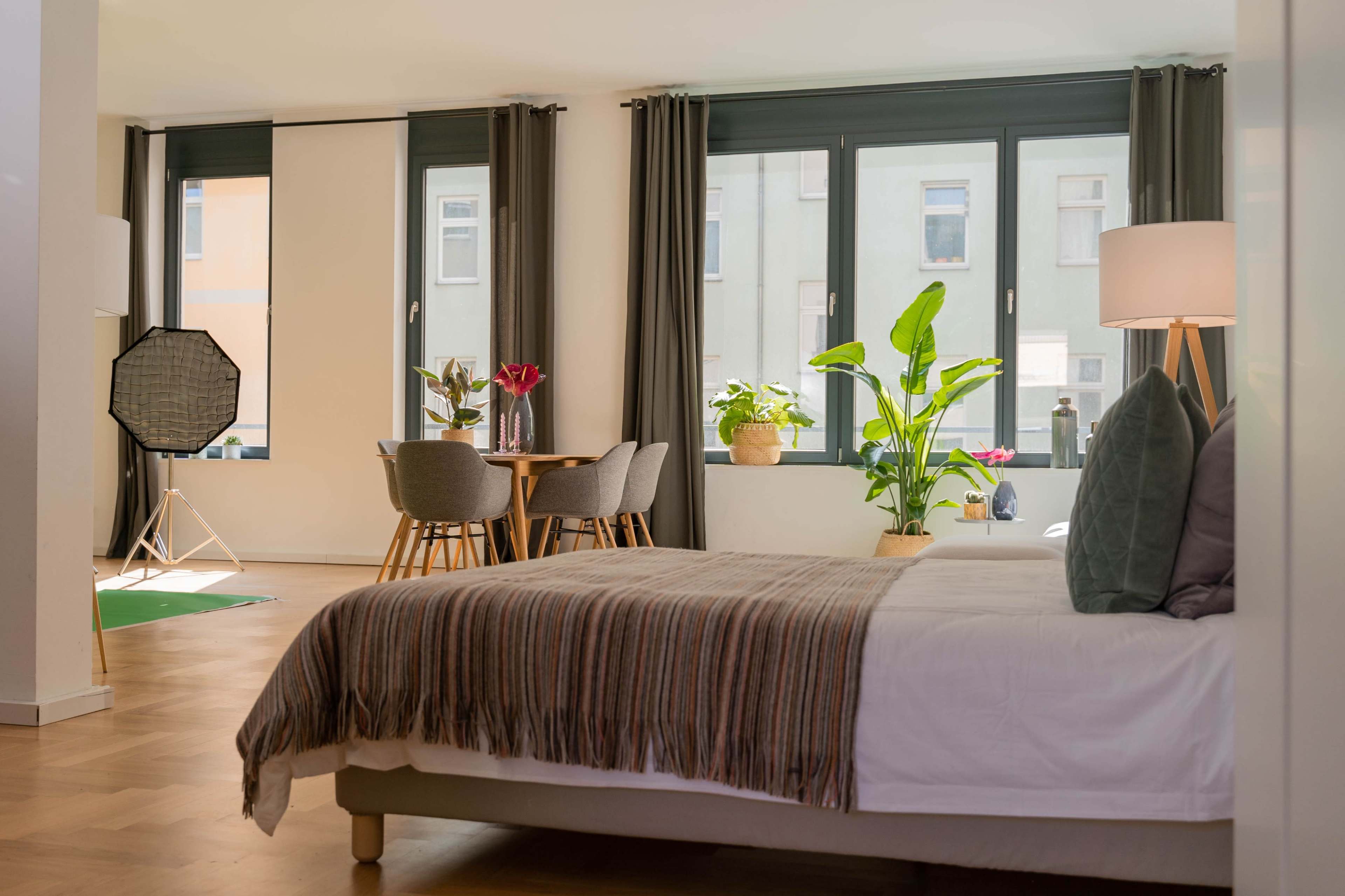 A modern bedroom features a bed with a throw, large windows, a dining area with chairs, and several potted plants.