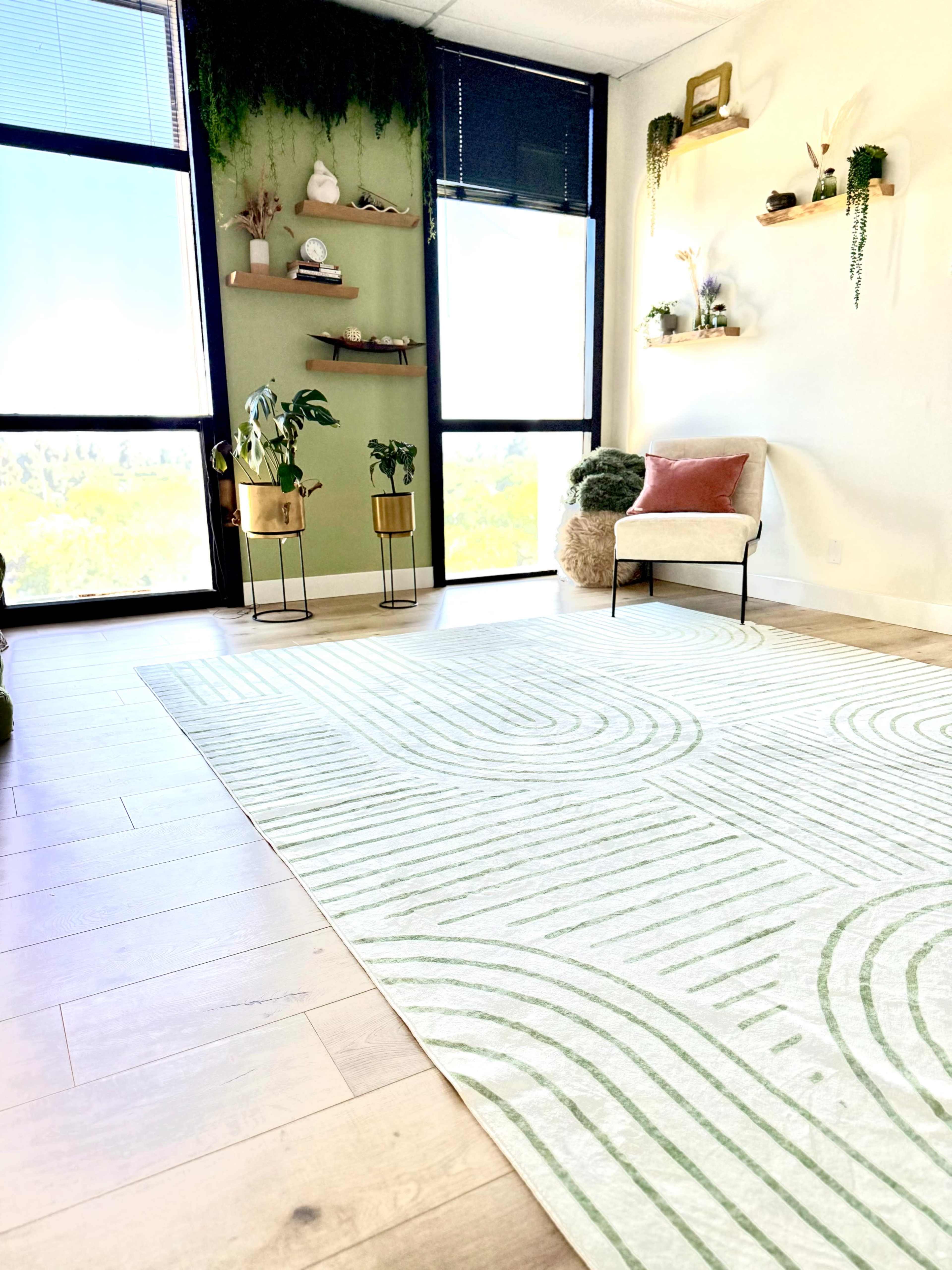 The image shows a bright room with large windows, a green accent wall, a decorative rug with curved lines, and a small seating area with a chair and cushions.