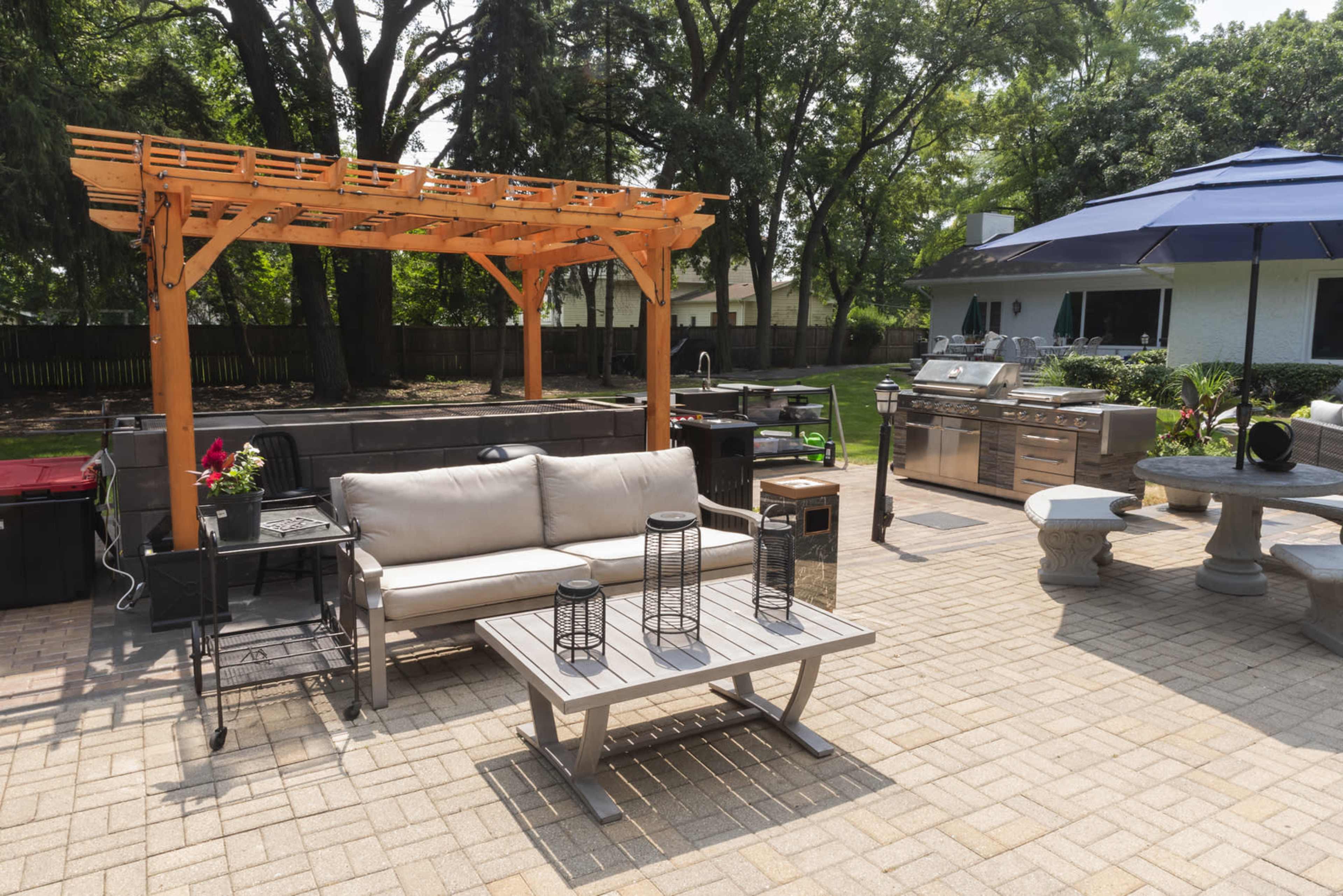 The image shows a patio area with a wooden pergola, outdoor furniture, and a grill, surrounded by greenery.