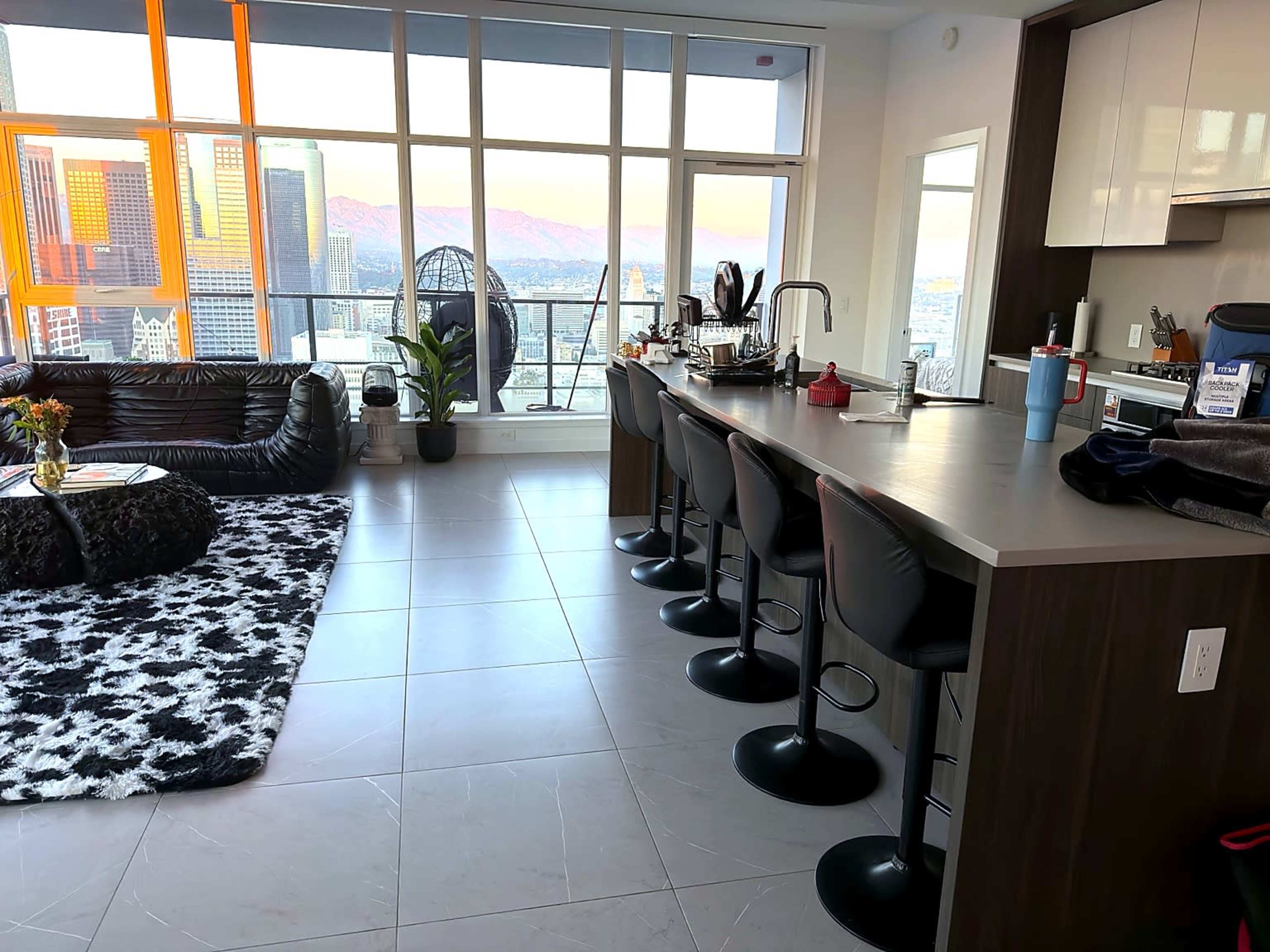 The image shows a modern kitchen and living area with a bar counter, several stools, and large windows offering a view of the city skyline and mountains in the background.